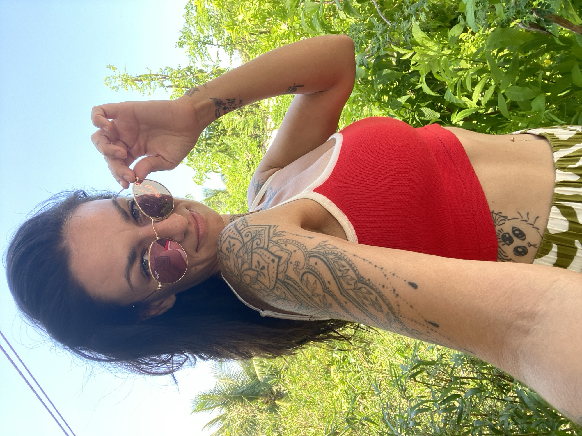 A woman with dark hair, wearing a red crop top, white and green striped pants, and sunglasses, poses for a selfie outdoors. Her left arm is adorned with intricate black tattoos. She is adjusting her sunglasses with her right hand. Behind her, lush green foliage and palm trees are visible against a bright blue sky. The scene suggests a tropical or subtropical location, consistent with Mũi Né, Vietnam.