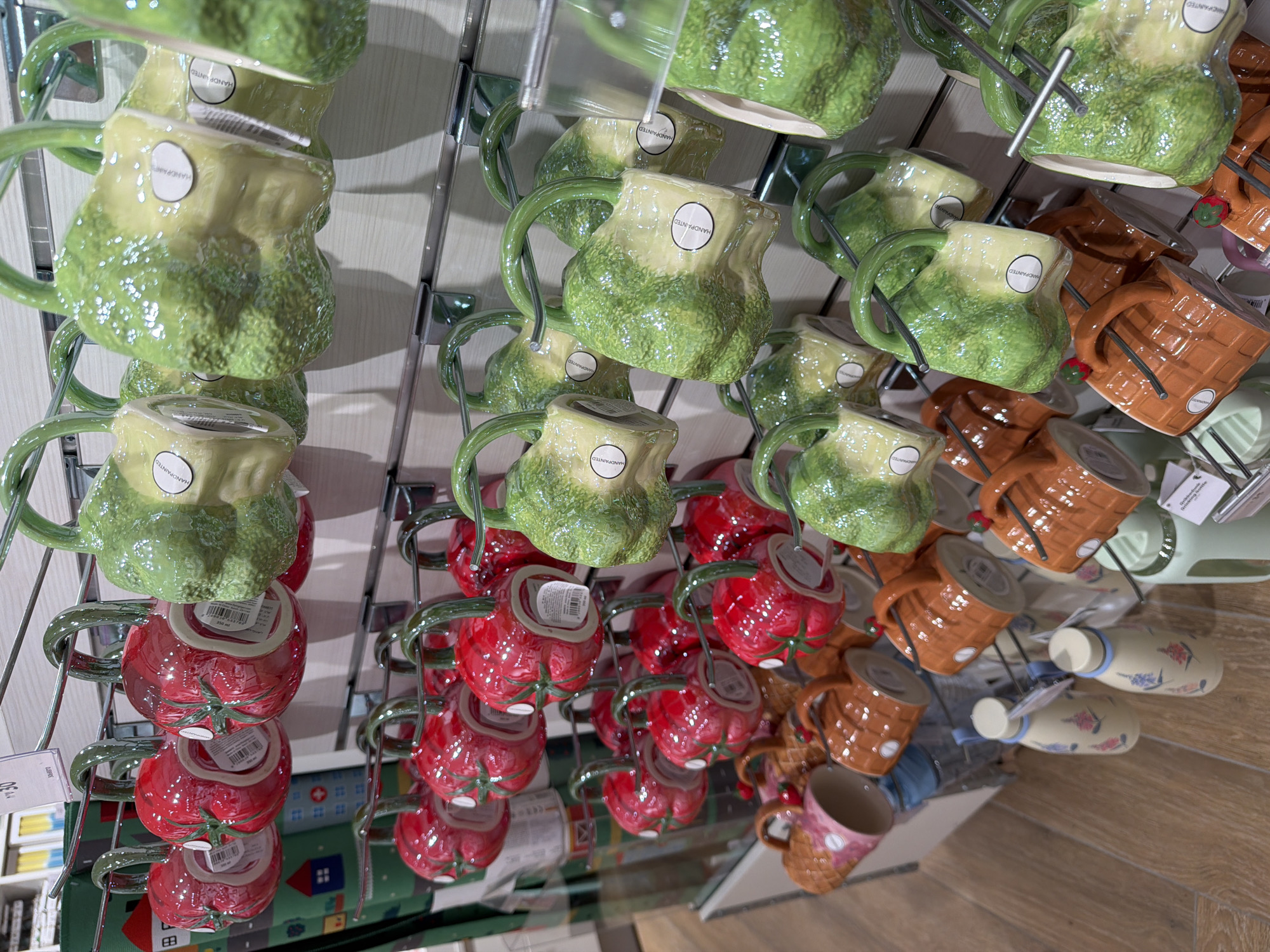 This image shows a display of mugs hanging on a metal rack in a store. The mugs are of various shapes and colors, including green, red (tomato-shaped), and brown (basket-weave pattern). Some mugs have a white label with the word "HANDPAINTED" on them. The mugs are likely for sale. The scene suggests a retail environment, possibly a home goods store. The city and state are not discernable from the image.
