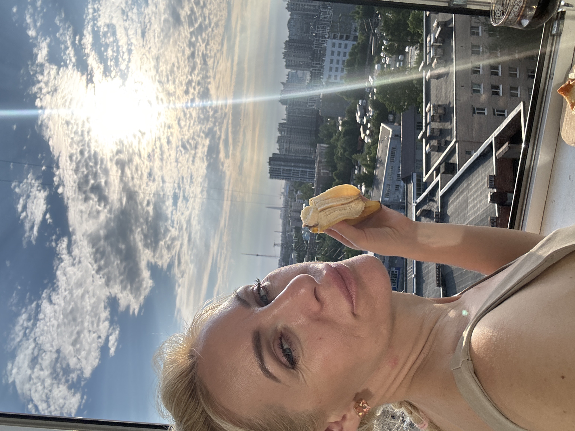 A blonde woman with light skin and blue eyes is holding a banana in her left hand and looking at the camera. She is wearing a light brown tank top and a square pink earring in her left ear. The background is a cityscape with tall buildings and trees under a cloudy blue sky with the sun shining through. There is also a glass of red wine and a plate with some food on a windowsill in the foreground. The overall scene appears to be a selfie taken from a high vantage point, possibly an apartment with a view.