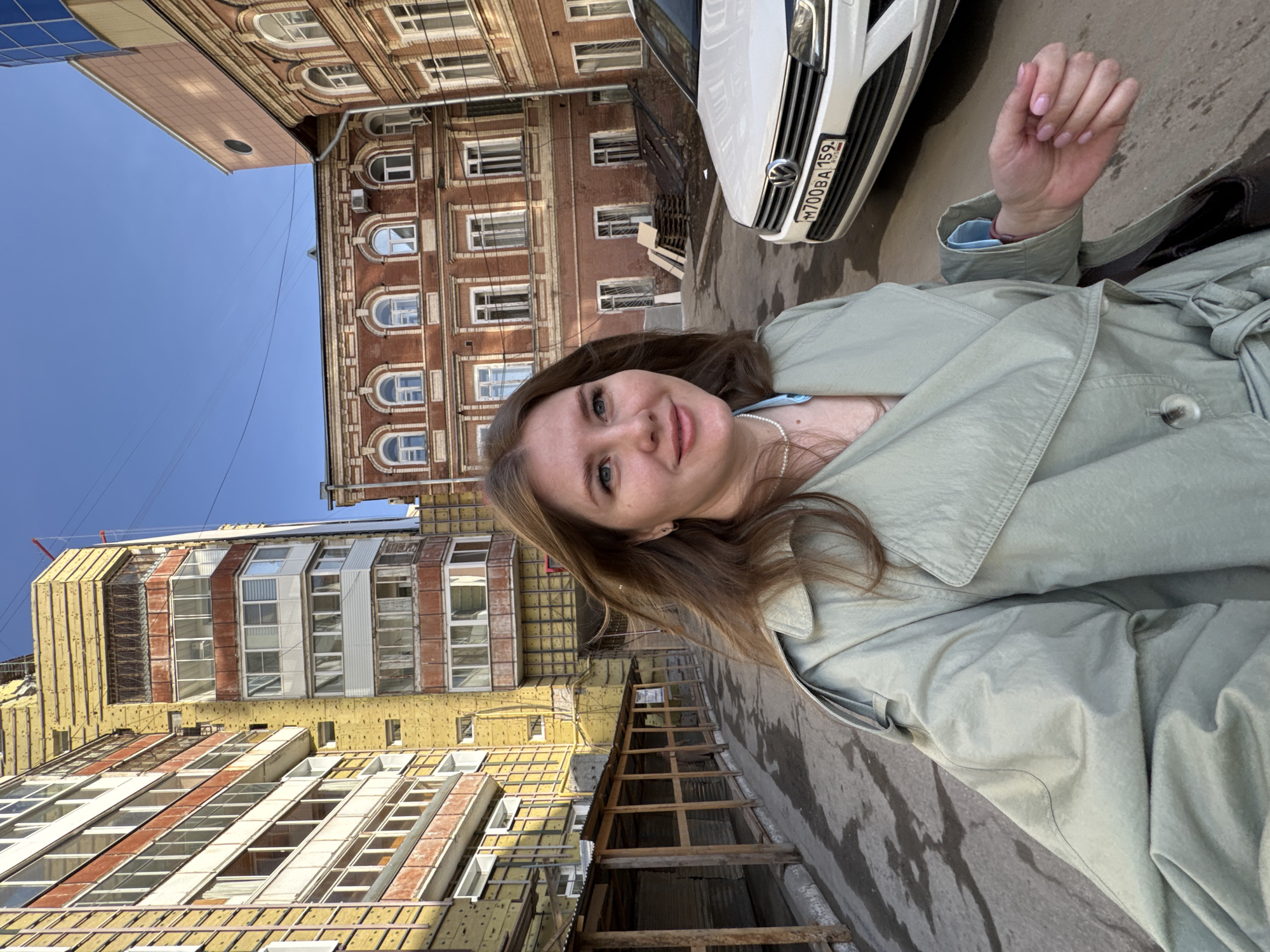 The image shows a young woman in a light green trench coat, standing on a sidewalk in Perm, Russia. A white Volkswagen car is parked beside her on the right, its license plate showing "M700BA 159 RUS." Behind the woman is a mix of old and new architecture. To the left is a partially renovated building with yellow insulation panels covering sections of its exterior. To the right is an older, red brick building with arched windows and detailed trim. The sky is a clear blue. The scene appears to be a public street in a city.