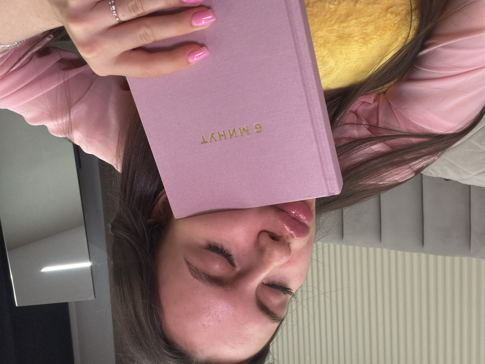 An indoor photograph, taken in the city of Chaikovsky, Russia, features a young adult female reclining. Her eyes are closed, and her lips are pursed in a puckered expression, coated with a glossy substance. She possesses long, dark brown hair and is clad in a pink long-sleeved garment. Her left hand, adorned with pink nail polish and a ring featuring three small stones on the ring finger, holds a pink hardback book against her face. The book's cover displays the title "6 МИНУТ" (6 MINUTES) in gold Cyrillic lettering. A yellow, textured object, consistent with a plush cushion or pillow, is partially visible beneath her. The background consists of a gray, vertically paneled upholstered headboard, behind which is a light-colored wall with thin vertical striations. To the right, a dark, reflective surface with a prominent horizontal light reflection is visible, indicating an indoor environment with artificial lighting.