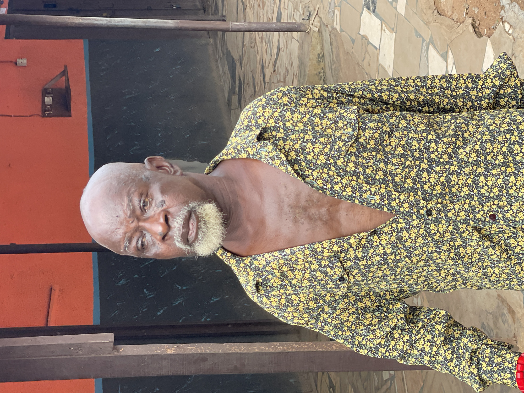 An elderly Black man is seen in Obosi, Nigeria. He has a bald head, a light beard, and wears a shirt patterned with small yellow flowers on a dark background. He stands near a building with a red and black wall, and a stone tile walkway. A metal beam runs horizontally in the foreground.