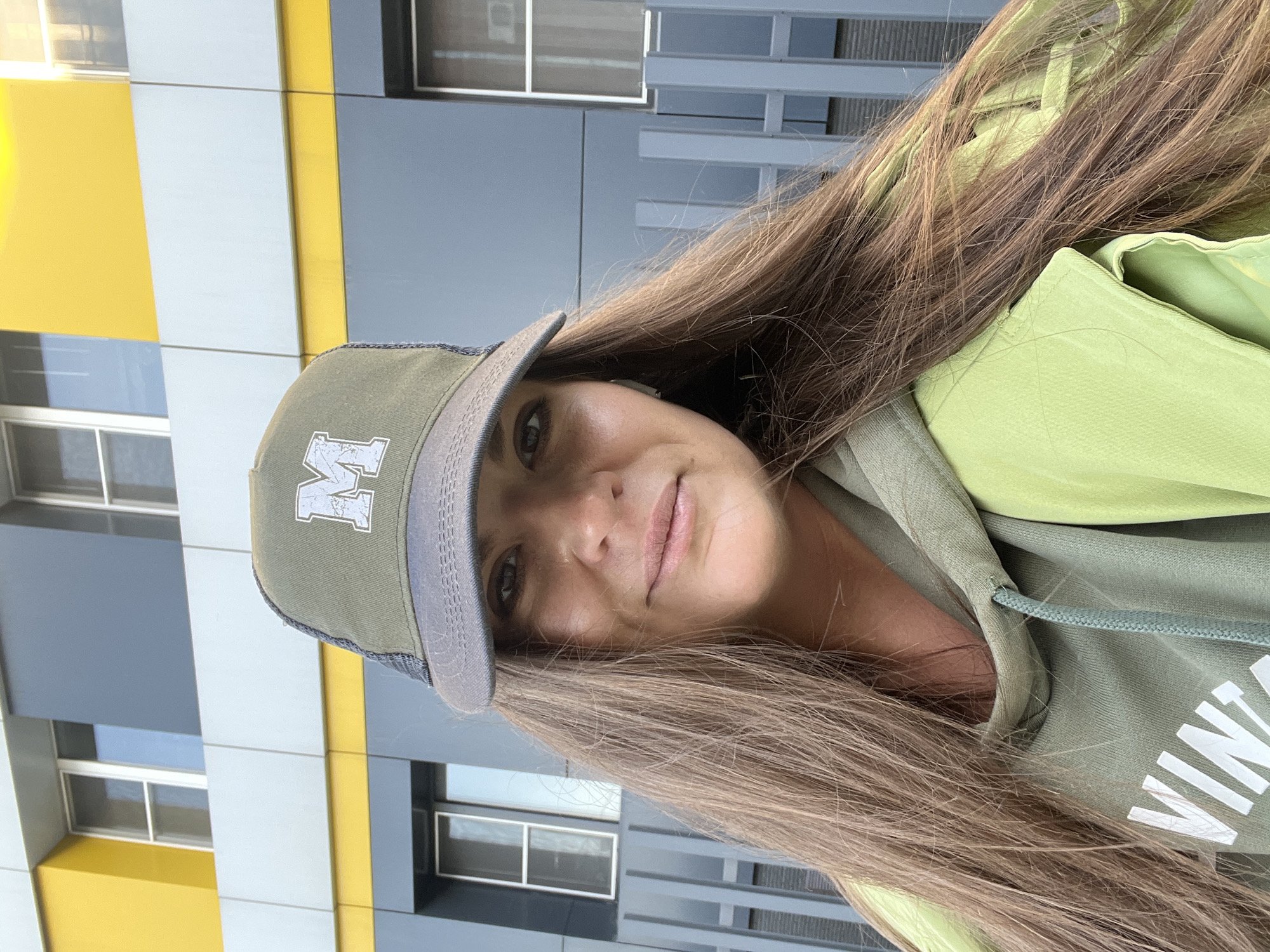A woman wearing a green baseball cap with a white "M" on the front and a green hoodie stands in front of a modern building with yellow, white, and gray panels. The building has windows visible in the background. The location is Novosibirsk, Russia.