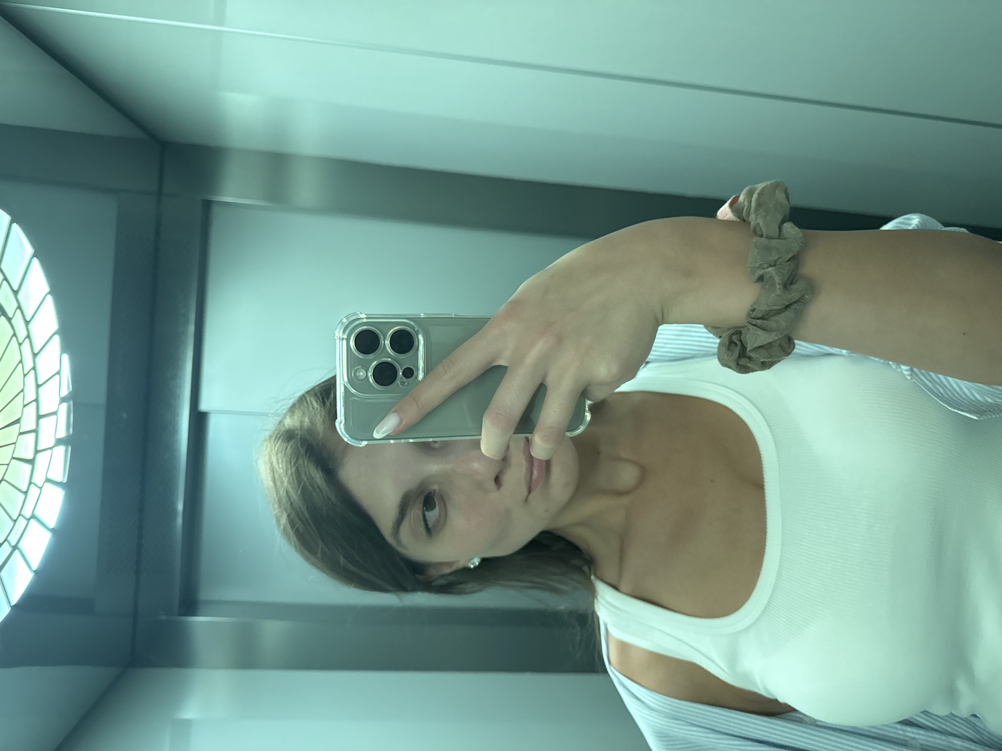 A young Caucasian woman with brown hair tied back and light makeup is taking a selfie in a mirrored elevator. She is wearing a white ribbed tank top and a light blue and white striped button-down shirt worn open. She has a scrunchie on her left wrist. The elevator has metallic walls and a tiled ceiling light fixture. The image does not provide any information about the city of Burgas, Bulgaria.