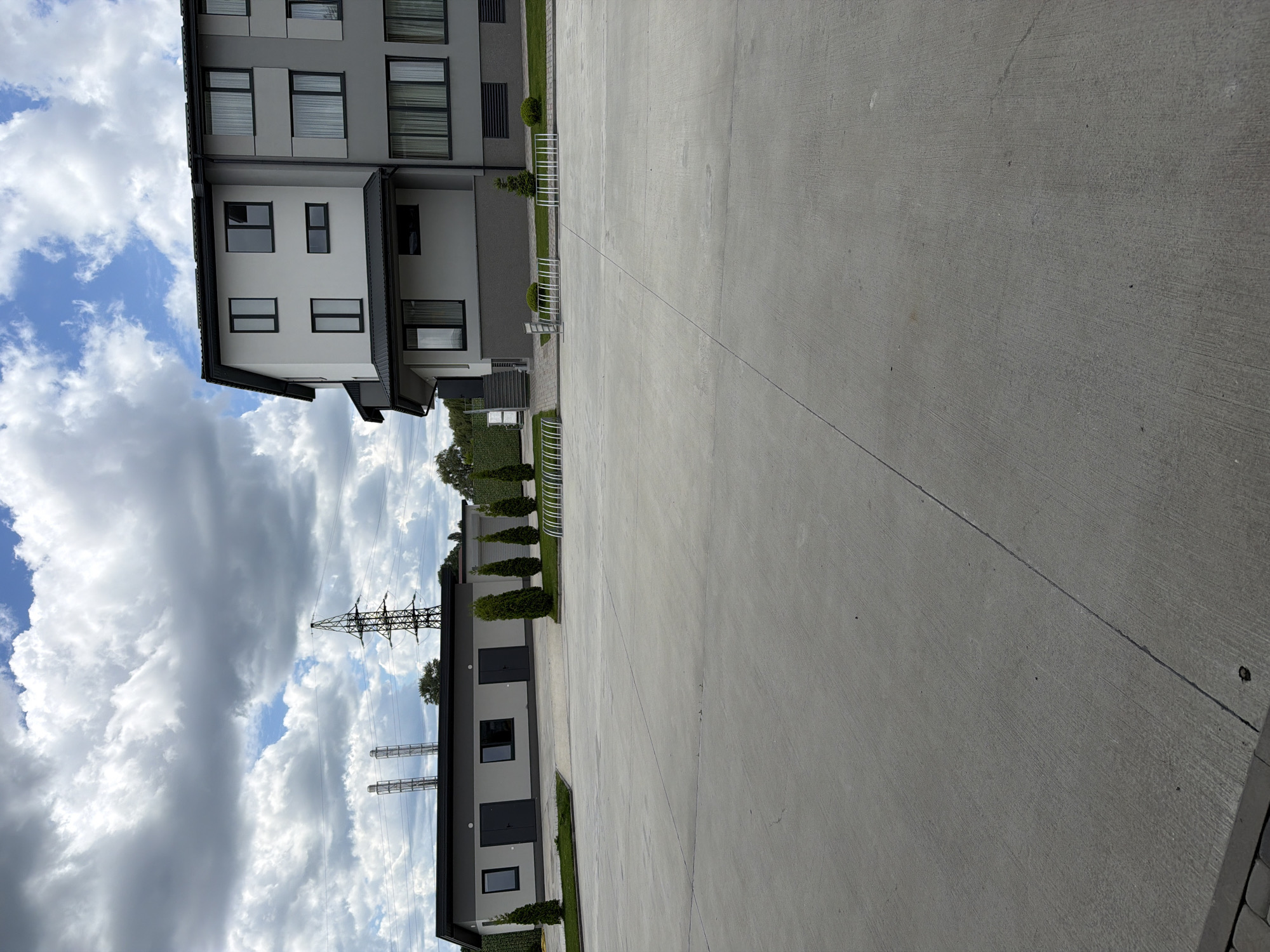 The image shows an outdoor paved area in front of a modern building complex. The building has a light gray and white facade with multiple windows. To the left of the main building is a smaller, single-story building with a dark roof and gray walls. The paved area appears to be a parking lot or courtyard. Several bicycle racks are visible in front of the buildings. The environment is characterized by a cloudy sky with patches of blue and a distant view of electrical pylons and trees. The scene is located in the city of Hnidyn, Ukraine. There are no people or animals visible in the image.