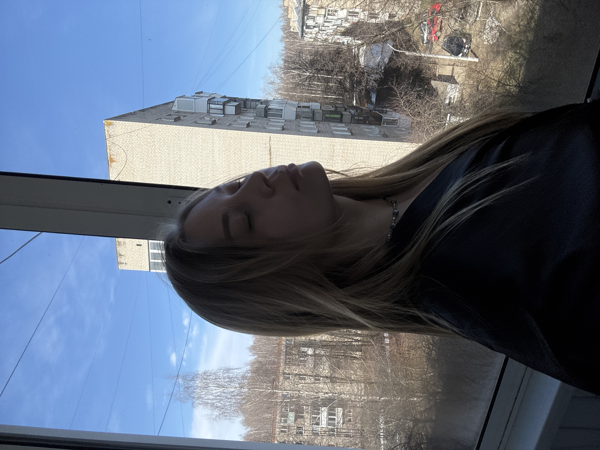 A woman with blonde hair is positioned near a window, with a view to the exterior. In the background, there is a building constructed from beige bricks, featuring multiple windows. Further out, a cluster of trees without leaves can be seen, as well as more buildings in the distance. The sky is predominantly blue. The woman is wearing a dark garment. The location is in the city of Chaikovsky, Russia.