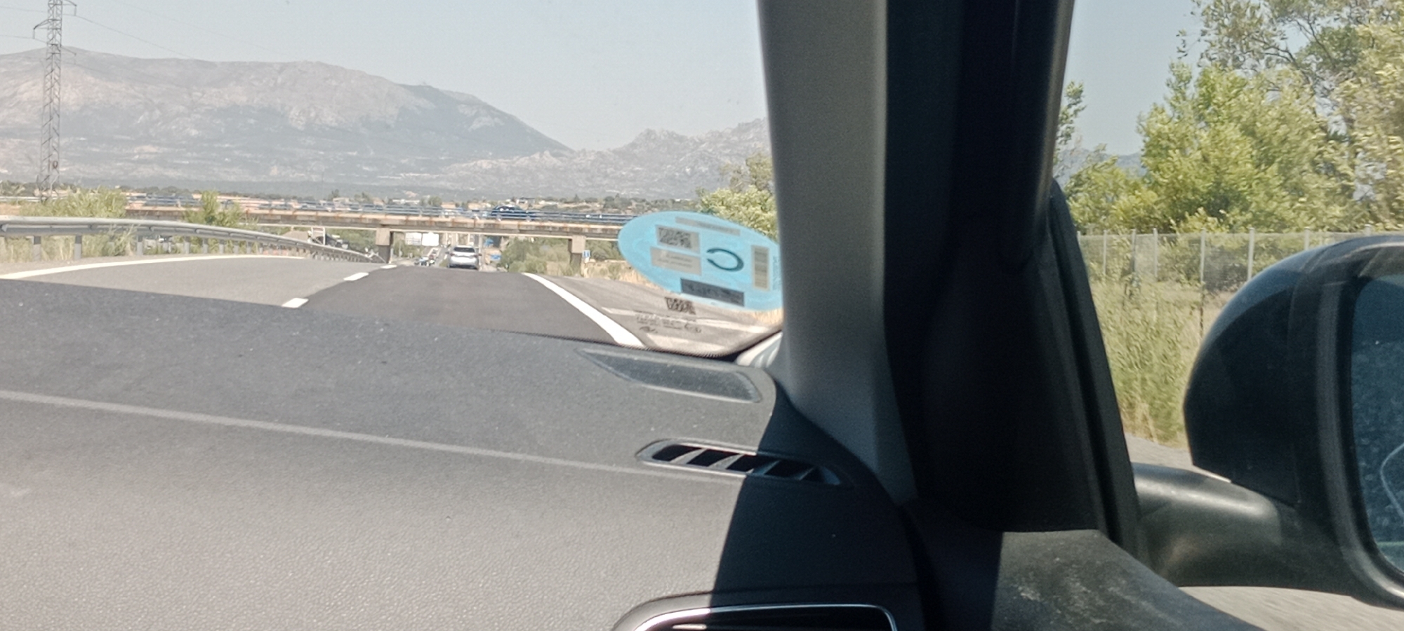 The image shows a view from the interior of a car driving on a highway near Pedrezuela, Spain. The road ahead curves to the left and is bordered by a guardrail on the left and a grassy embankment on the right. In the distance, there is a bridge structure spanning the highway, with traffic visible on it. Beyond the bridge, a mountain range is visible under a clear sky. A white car is driving ahead in the same lane. On the dashboard of the car, a blue sticker with circular and rectangular elements is affixed to the windshield. The right side of the car's interior, including the passenger side pillar and a portion of the side mirror, is also visible. The overall scene depicts a typical highway driving environment on a sunny day.
