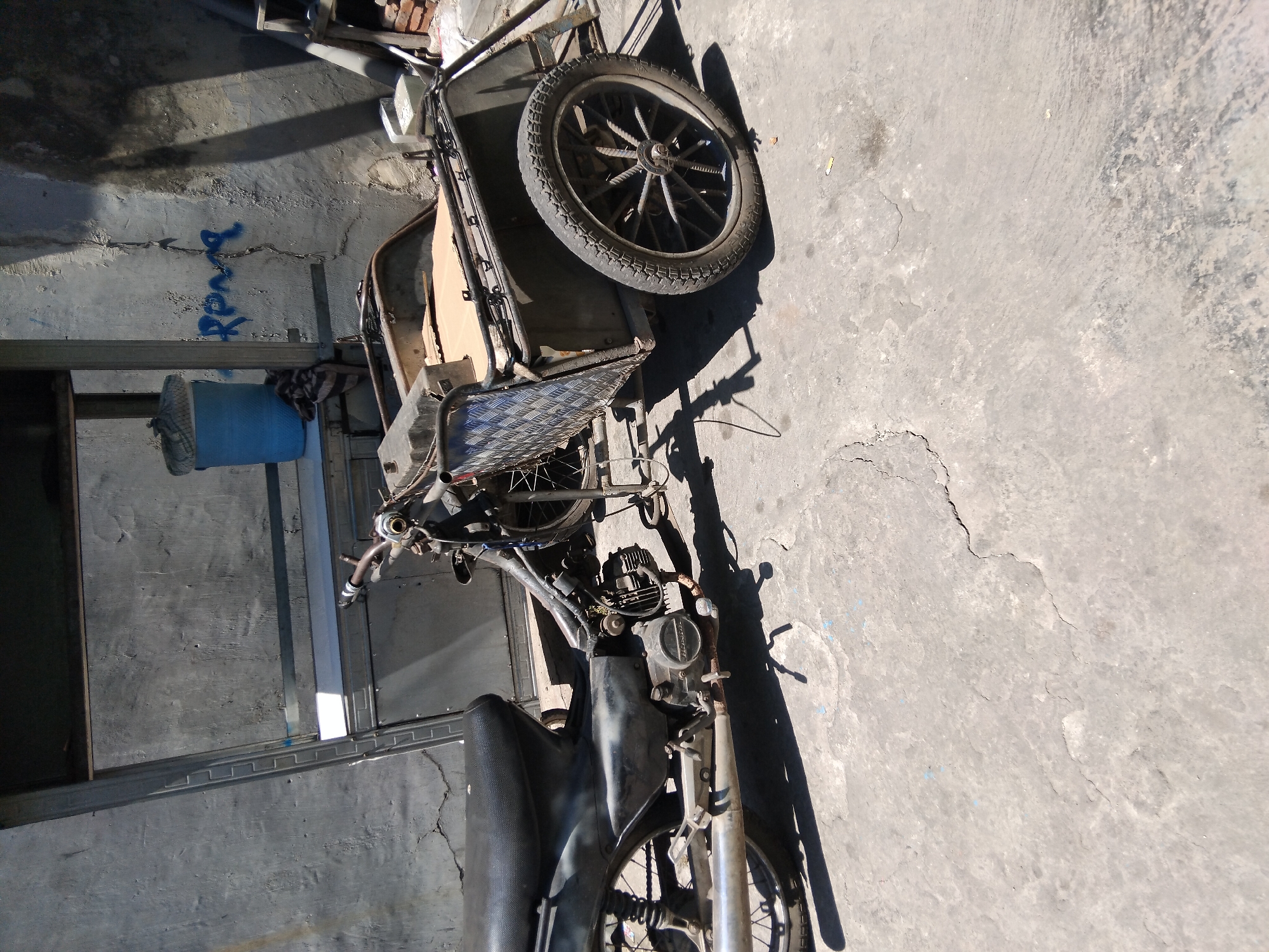 A weathered motorcycle with a sidecar is parked on a cracked concrete surface, casting shadows from the harsh sunlight. The motorcycle is dark and appears to be a utility vehicle, possibly used for transport or work. The sidecar is a box-like structure with a metal diamond plate on its side, suggesting durability. In the background, a building with a textured wall is visible, with some blue graffiti scrawled across it. A blue bucket and some fabric are also present near the building. The overall scene depicts an outdoor area, likely in a residential or commercial space in Kepuh Kiriman, Indonesia, given the context. The environment is utilitarian and shows signs of wear and tear.