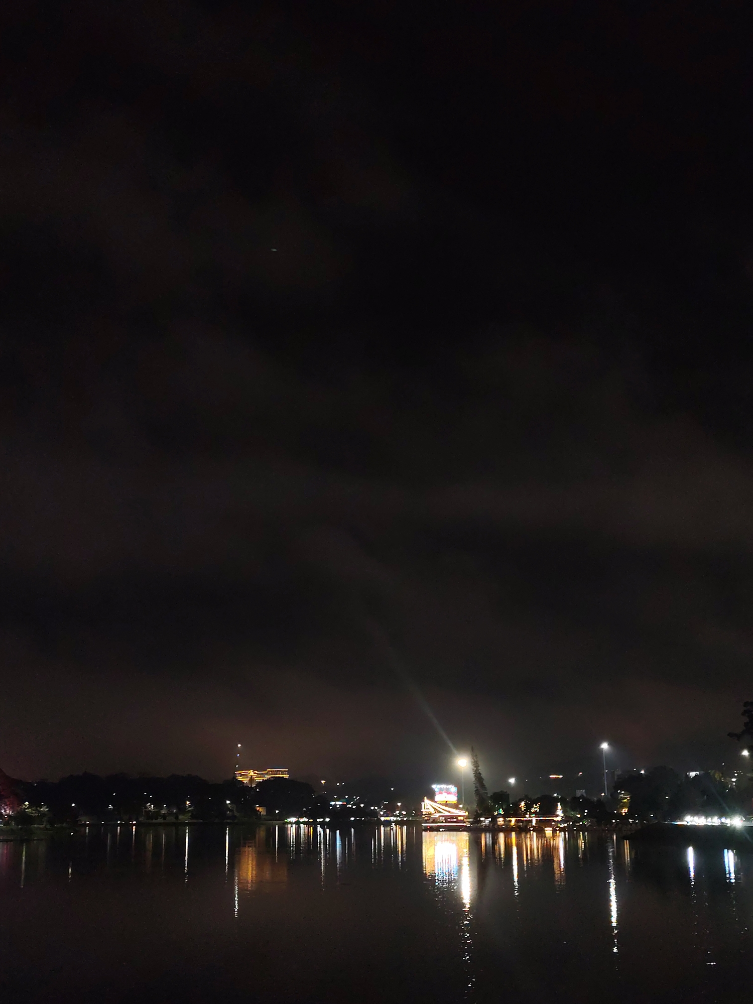 The image depicts a night scene of Ho Xuan Huong Lake in Da Lat, Vietnam. The lake reflects lights from buildings and streetlights along the shoreline. The sky is dark and cloudy. The lights create vertical reflections on the water surface. Trees and vegetation are visible along the lake shore. There are no people visible in the image.