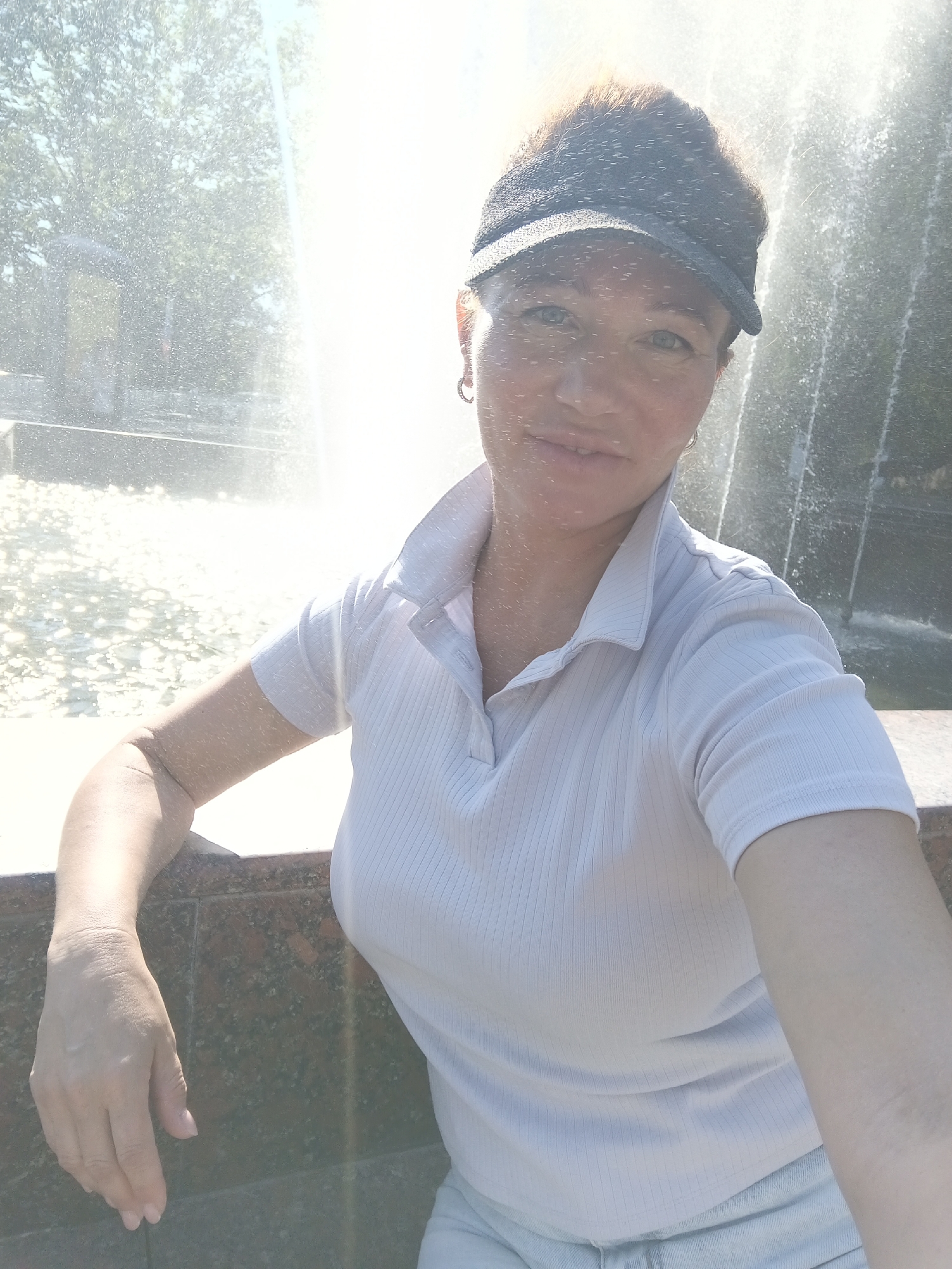 A medium-shot, selfie-style photograph features a woman in her late 30s or early 40s, with reddish-brown hair styled into a messy bun and a dark baseball cap, posing in front of a bright, sunlit fountain. The woman is wearing a light blue, ribbed polo shirt and appears to be outdoors in a sunny environment, suggested by the bright light and water spray. Her left arm is bent and resting on a dark, textured ledge, possibly marble or granite, that frames the lower part of the image. The fountain behind her is active, with water cascading down and creating a bright, blurred background, indicative of a public park or recreational area. The overall impression is one of a casual, outdoor moment captured on a warm day. Based on the visible fountain and potential park setting, this scene could be located in a city like Sochi, Russia, known for its numerous parks and fountains.