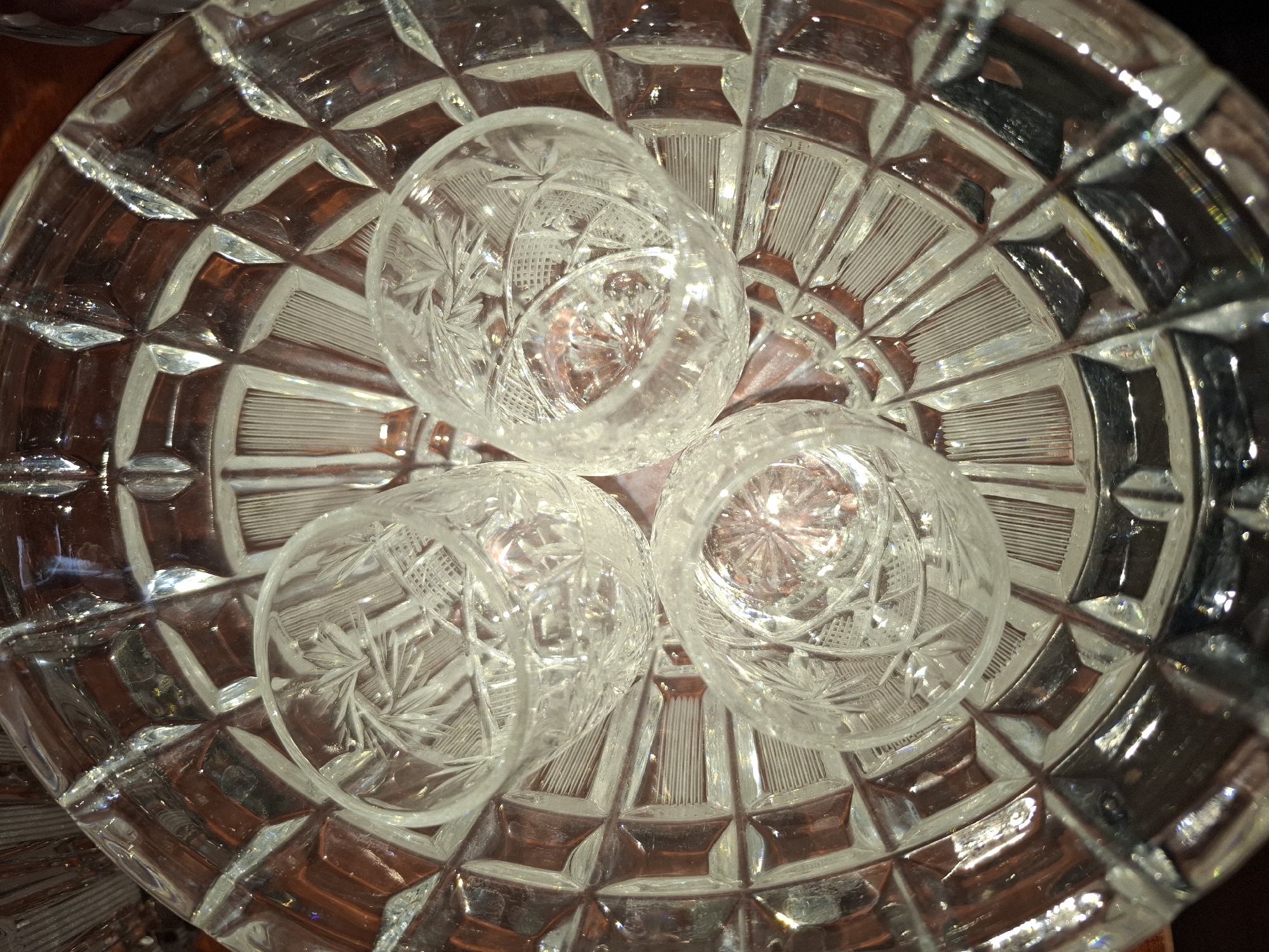 The image displays a close-up, overhead view of three small, intricately patterned crystal shot glasses nestled within a larger, faceted crystal bowl. The crystal pieces exhibit detailed starburst and geometric etchings, catching and reflecting light. The larger bowl has a diamond-shaped pattern with radiating lines. The background appears to be a dark, possibly wooden surface. The scene is static and lacks any human presence. The image does not provide any information about the city of Almaty, Kazakhstan.