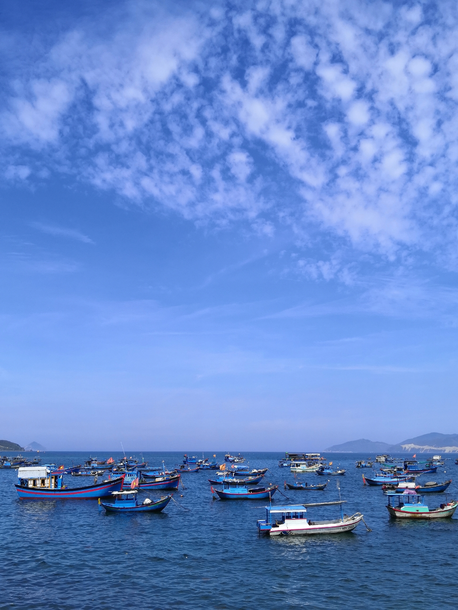 A wide-angle, high-angle shot depicts a fleet of colorful fishing boats bobbing on the blue ocean near the coast of Bac Nha Trang Ward, Vietnam. The sky above is a vibrant blue, dotted with scattered white clouds, hinting at a clear and pleasant day. The boats, primarily in shades of blue, red, and white, are moored together, creating a dense collection on the water's surface. Some boats appear to be larger vessels with cabins and multiple decks, while others are smaller, more open fishing boats. The water itself is a deep, rich blue, with gentle ripples indicating calm conditions. In the background, the coastline is visible, with low-lying hills or mountains receding into the distance under the bright sky. The image captures a typical scene of maritime activity in a coastal Vietnamese community.