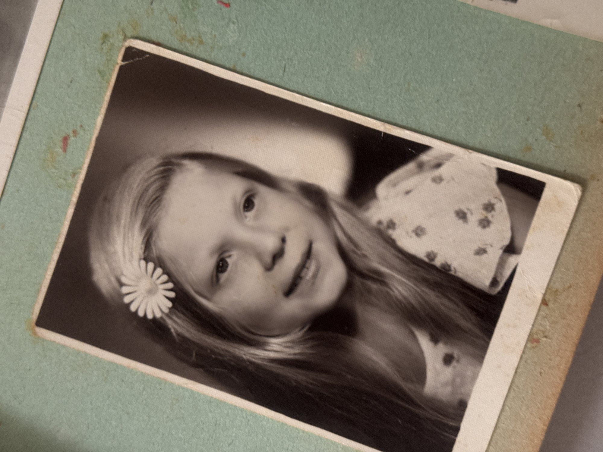 A slightly askew, close-up shot depicts a black and white photograph resting on a light green, textured surface, likely a page in a photo album. The photograph portrays a young girl with long, light-colored hair adorned with a white daisy tucked behind her ear. She is wearing a light-colored dress with a small, repeating floral pattern. The album page shows some wear with stains and discoloration in the corners. The photograph itself appears to be old with signs of age, such as slight fading. There is no evidence to suggest that this image was taken or found in Ramat Gan, Israel.