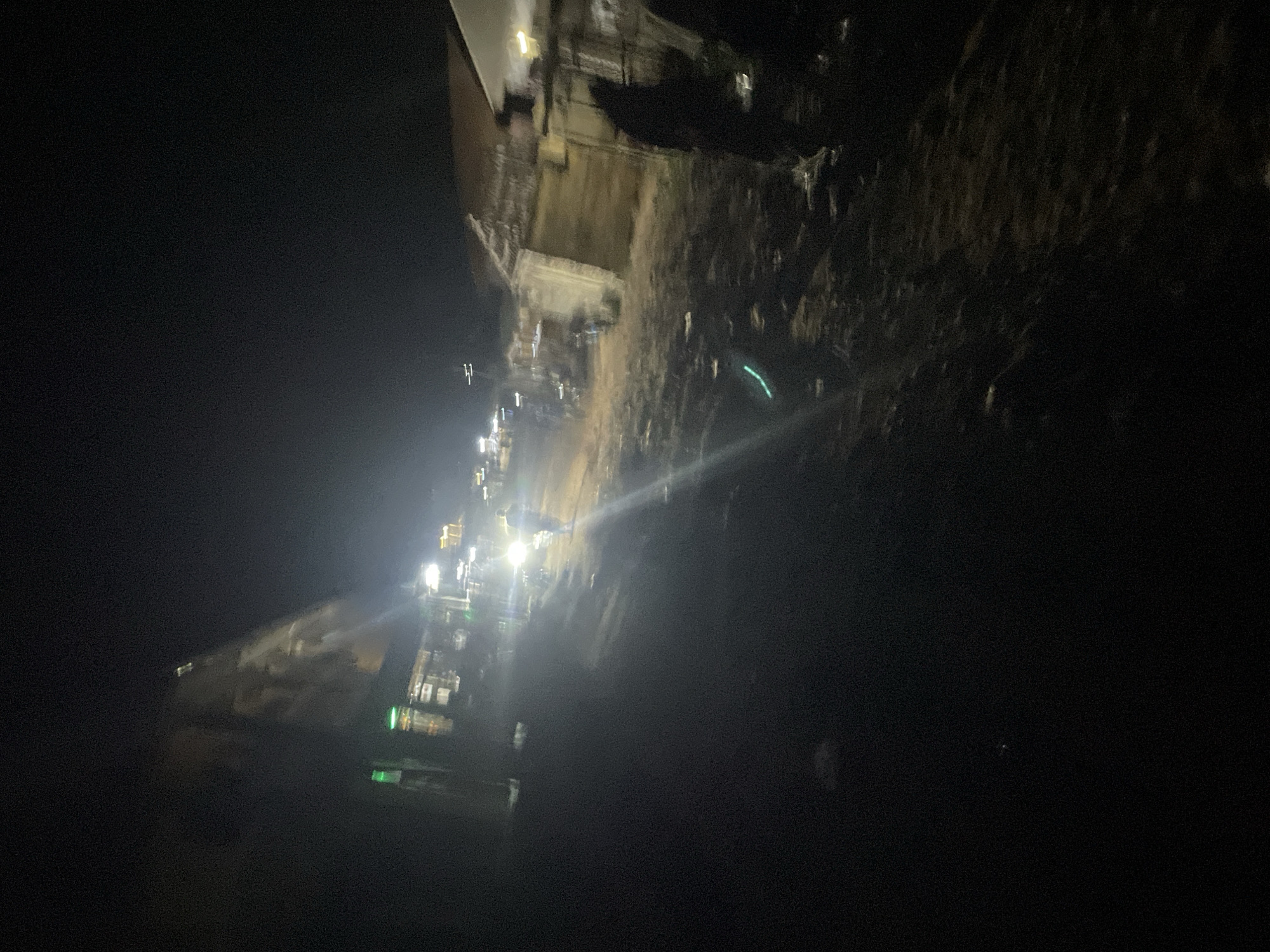The image depicts a dimly lit street scene at night in Lokoja, Nigeria. A prominent structure, appearing to be a building or a large advertisement, is visible on the left, casting a faint glow. Several smaller buildings with illuminated windows line the street on the left side, suggesting commercial activity. In the center of the image, a person is walking away from the camera, illuminated by a bright headlight from an unseen vehicle. The street surface appears to be unpaved or dirt. On the right side of the street, more buildings are visible, with one structure featuring a corrugated roof and another appearing to be a wall. Shadows are cast across the ground, indicating the presence of artificial lighting. The overall atmosphere is one of a modest, possibly rural or semi-urban, setting at night.