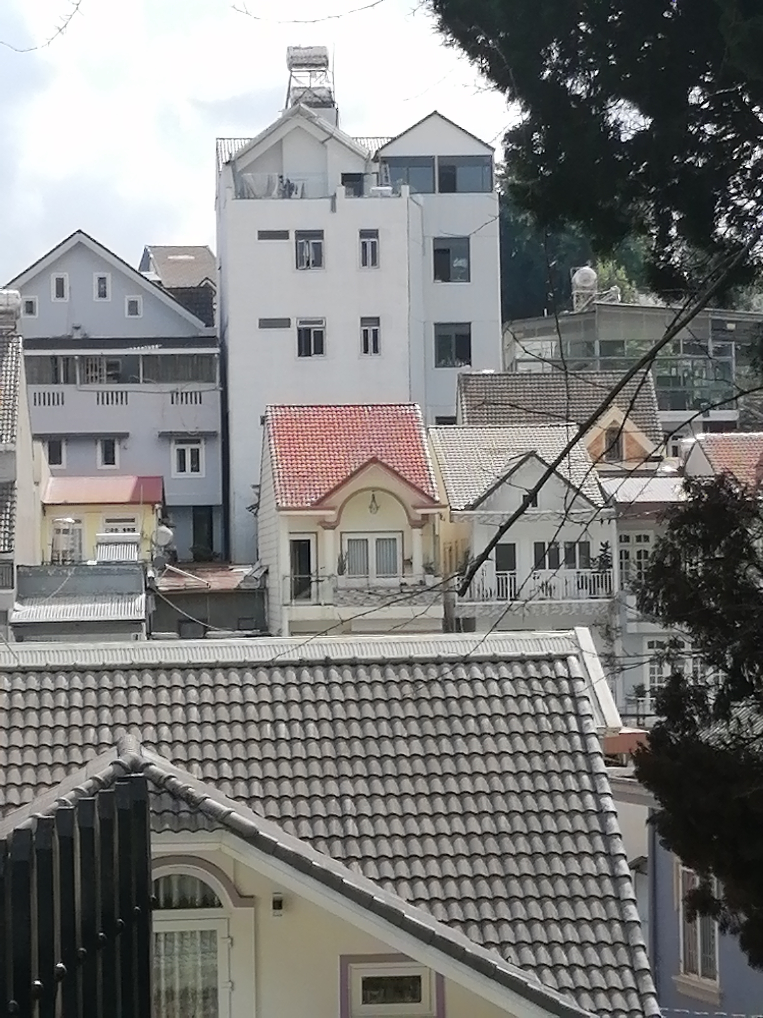 This is a daytime, eye-level, full shot of a dense urban hillside in Phường Xuân Hương, Đà Lạt, Vietnam. The scene is dominated by multi-story buildings with predominantly white and light blue facades. The architecture is varied, with some buildings featuring modern, flat roofs and others with classic tiled, pitched roofs. Many windows, some with shutters, are visible. On the rooftop of one of the tallest buildings, a solar water heater is visible. The foreground is occupied by the tiled roof of a lower building and a section of black metal fencing. In the background, lush green trees frame the view, suggesting the hilly terrain of Đà Lạt. The sky is overcast with scattered clouds. There are no people or animals clearly visible in the image.
