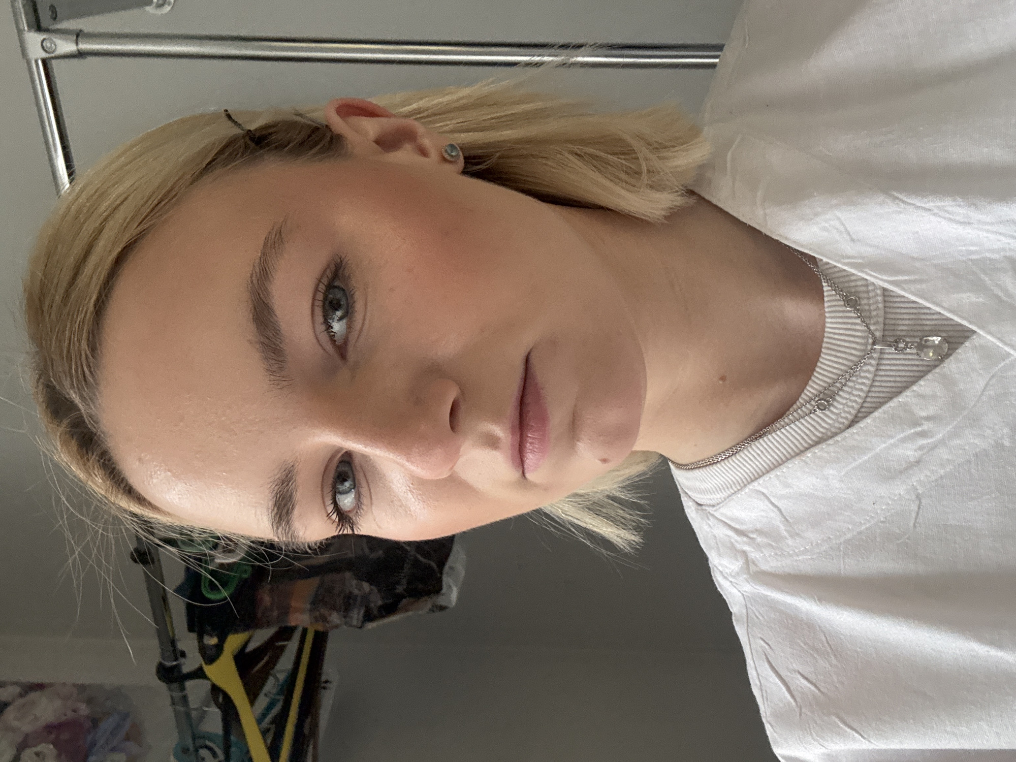 A close-up portrait of a light-skinned woman with blonde hair and blue eyes is captured. She is wearing a white t-shirt and a delicate silver necklace with a small pendant. A small earring is visible in her left ear. A small black clip can be seen in her hair. In the background, there is a glimpse of a clothing rack with various items. There is no indication that this image was captured in St. Petersburg, Russia.
