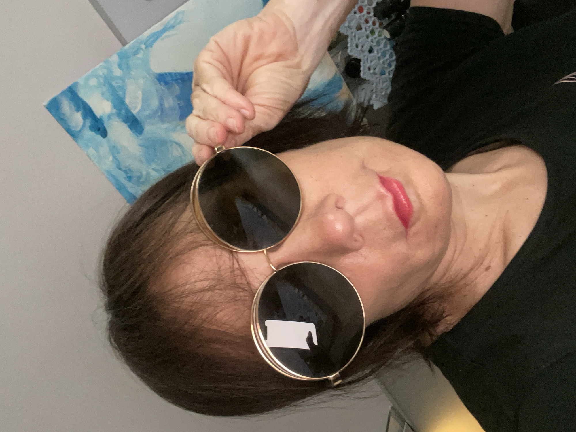 A medium shot captures a woman wearing large, round, gold-rimmed sunglasses and a black t-shirt. She has dark brown hair with bangs and is holding the edge of her sunglasses with her right hand. Her lips are painted a bright pink. A painting with abstract blue and white brushstrokes is visible in the background, along with a white lace-like object. The scene appears to be indoors. The city of Sochi, Russia, is not discernible in the image.