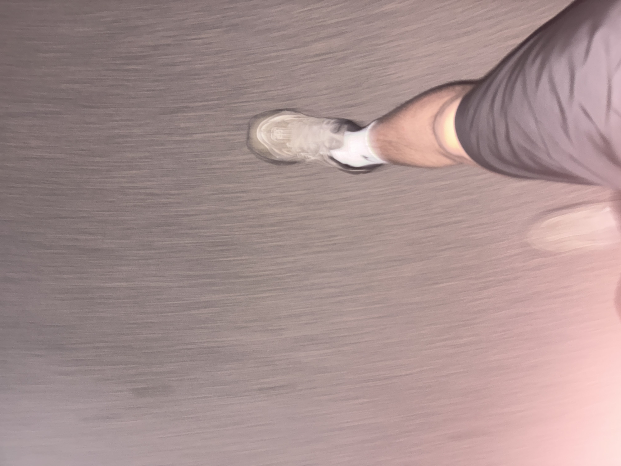 The image provides a close-up, top-down perspective of a person's right leg and foot. The subject is wearing dark grey or black athletic shorts, a white sock, and a light-colored (likely white or off-white) athletic shoe with a substantial sole. The skin on the leg is visible, appearing to be a light tone.
The scene depicts motion, as evidenced by the significant horizontal motion blur across the entire background. This blur suggests the person is actively walking or running. The ground surface is indistinct due to the blur, but appears to be a uniform grey-brown, possibly a paved surface, a road, or a treadmill.
A notable detail is a soft, reddish-pink light source or reflection visible in the bottom right corner of the image, casting a warm glow onto the ground and the person's leg in that area. The overall lighting is somewhat dim, which, combined with the reddish glow, could indicate an indoor setting, evening, or night conditions. No specific location cues like landmarks or weather elements are discernible. There is no visible text on the clothing or footwear.