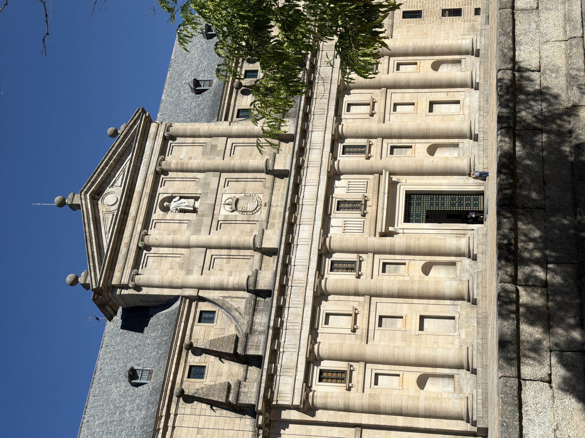 This is a photograph of the Monastery of San Lorenzo de El Escorial in San Lorenzo de El Escorial, Spain. The building is a large, symmetrical Renaissance structure made of tan stone. It features a prominent central facade with columns, a pediment, and statues. The roof is gray and tiled, with dormer windows. In front of the building, there is a stone plaza and a low stone wall. A few people are visible on the plaza, and a tree with green leaves is in the foreground on the right side of the image. The sky is clear and blue.