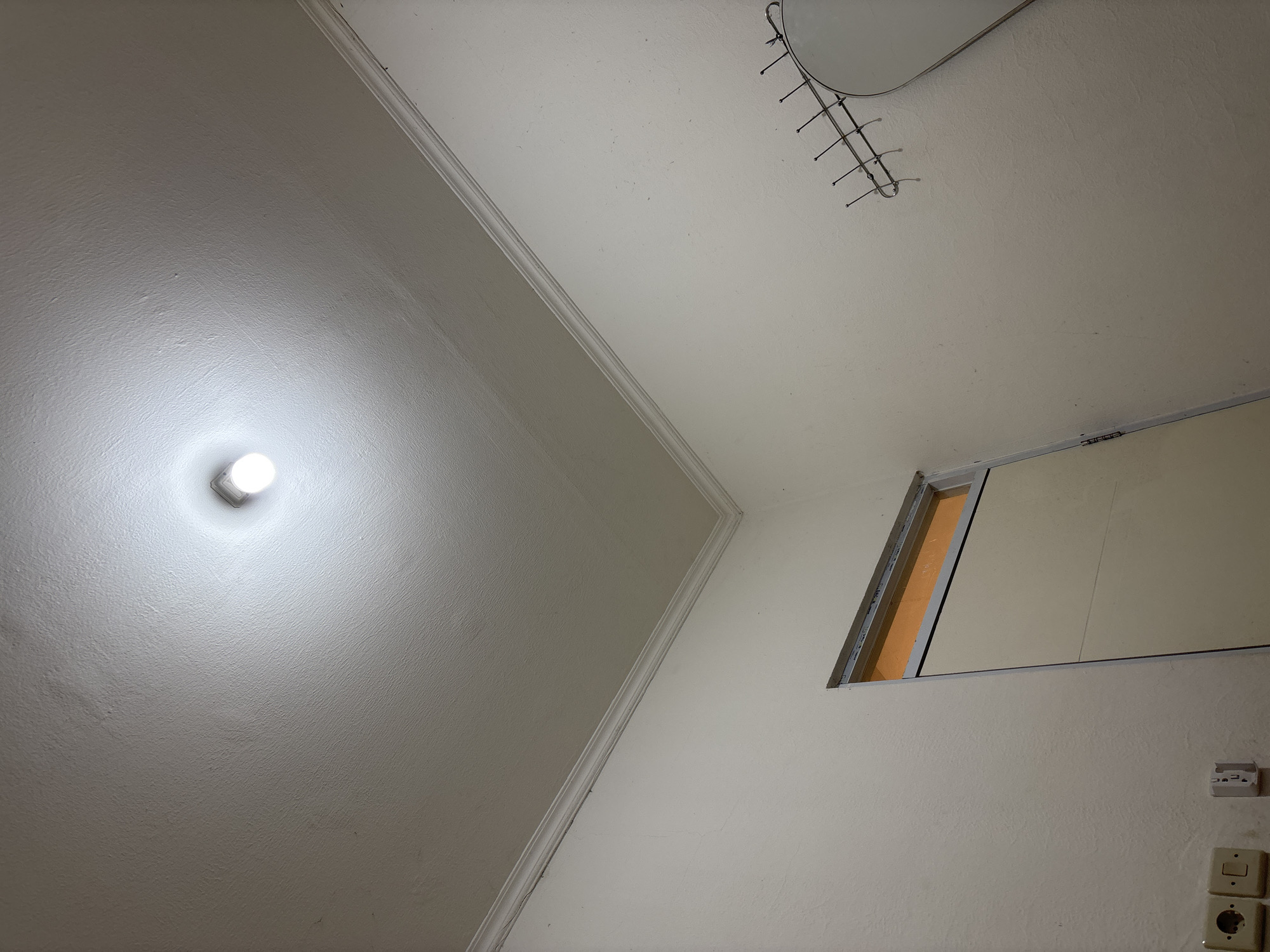 The image depicts an interior corner of a room, likely in a residential setting in Wanajaya, Indonesia. The ceiling is textured and painted white, with a single, bright light bulb illuminated in the center. The walls are also painted white and appear to have a slightly textured finish. A white decorative molding runs along the junction of the ceiling and the walls.
On the left wall, there are two electrical outlets and a light switch. Below the light switch, there is a partially visible door or cabinet with a beige surface and a silver frame. The upper portion of this opening shows a glimpse of an orange-tinted interior.
On the right wall, there is a metal coat rack with several hooks. A portion of a large, oval mirror is visible on the far right of the image, reflecting the light from the ceiling. The overall impression is one of a simple, functional interior space. There are no people or animals visible in the image.