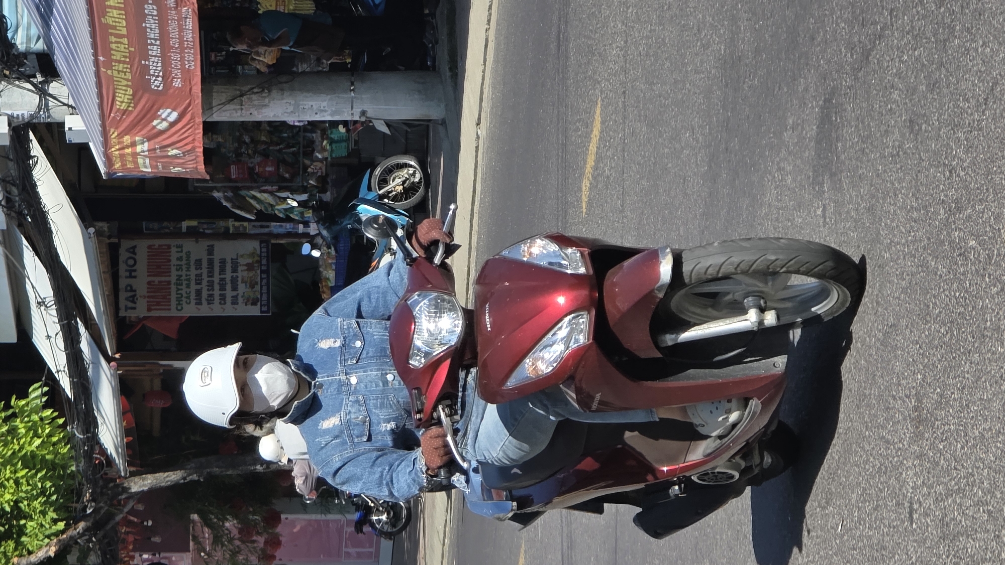 The image depicts a person on a red Honda scooter, wearing a white helmet, a face mask, a denim jacket, and gloves. The individual is riding on a paved road. The scooter is positioned diagonally across the frame. In the background, there is a storefront with a sign in Vietnamese that includes the words "TẠP HÓA THẲNG NHUNG" and lists various items sold in the store. There is another sign advertising sales, indicating it will take place over two days. Based on the signs and the scooter brand, the image is likely taken in the city of Nha Trang, Vietnam. Additional parked motorbikes can be seen in the vicinity of the storefront. Electrical wires are also visible above the store.