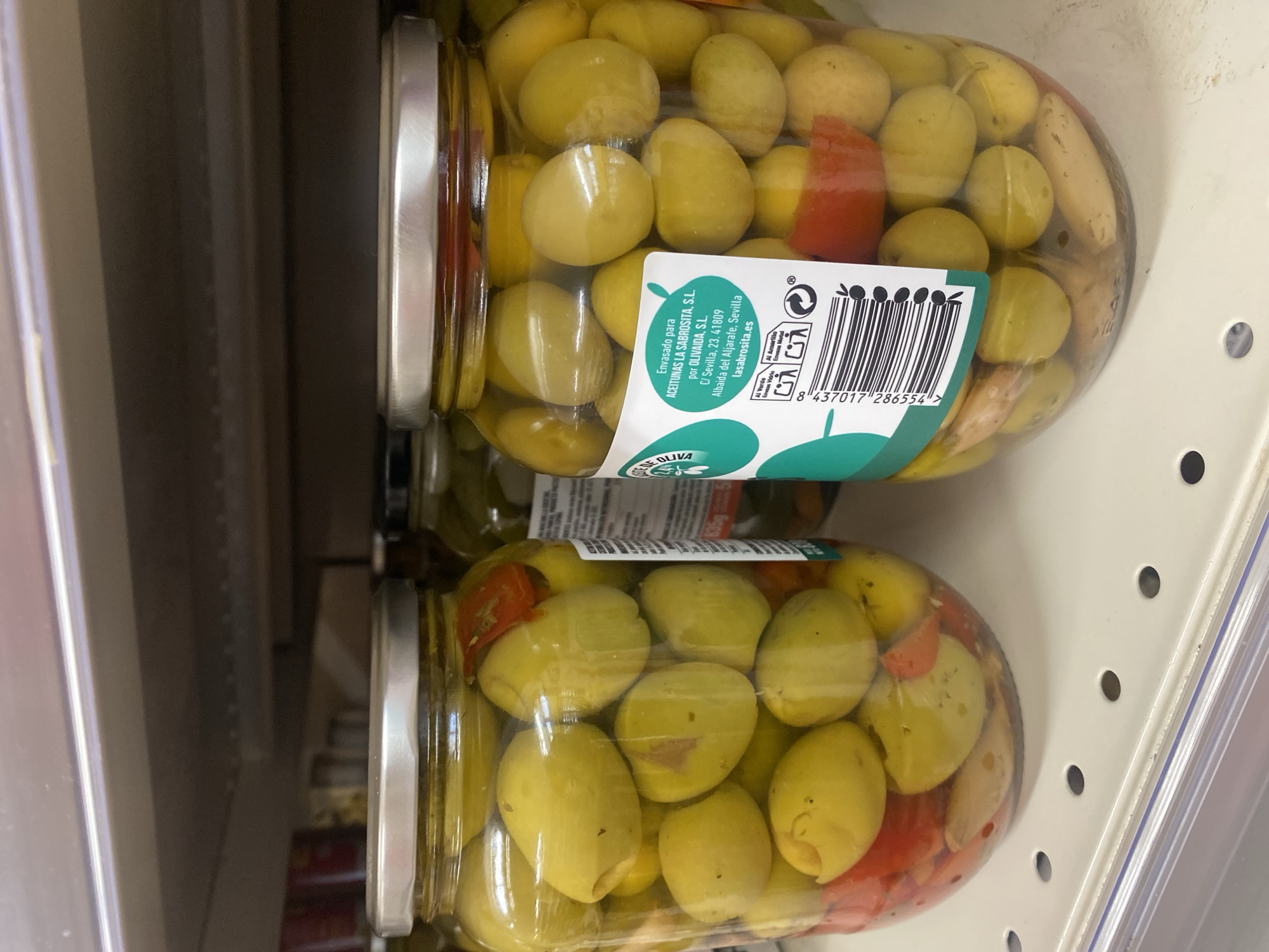 The image shows two glass jars of green olives with red bell pepper pieces, displayed on a shelf in a grocery store. The jars are clear, allowing the contents to be fully visible. The olives are plump and appear to be in brine. The top jar has a label with Spanish text and a barcode. The label reads "Envasado para ACEITUNAS LA SABROSITA, S.L. por OLIVAIDA, S.L. C/ Sevilla, 23. 41809 Albaida del Aljarafe, Sevilla lasabrosita.es" and "DE OLIVA". Below this, a barcode is visible along with the number "8 437017 286554 >". The scene is brightly lit, suggesting an indoor retail environment. The setting appears to be a supermarket aisle, with shelves and other products potentially out of frame. The focus is on the jars of olives, highlighting their contents and packaging. No people are visible in the image. The overall impression is a product shot of olives for sale.