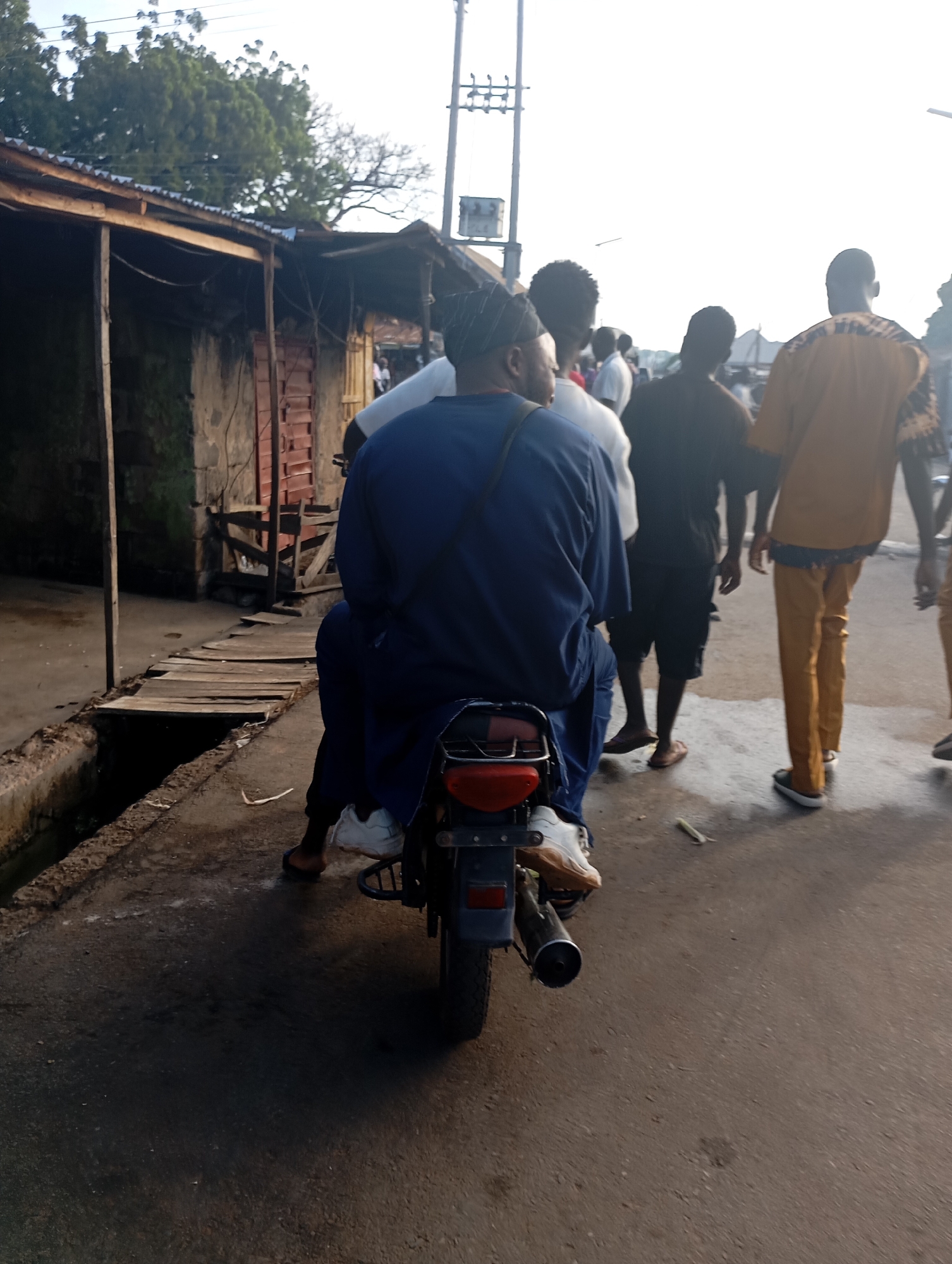 In Shendam, Nigeria, a man wearing a blue traditional outfit and a patterned cap rides a motorcycle, with another person seated behind him. The motorcycle is positioned on a dirt road next to a gutter and a wooden walkway that leads to a rustic, open-air structure with a corrugated metal roof. Several other individuals, dressed in casual attire and traditional Nigerian clothing, are visible in the background. The sky is bright, suggesting daylight hours.