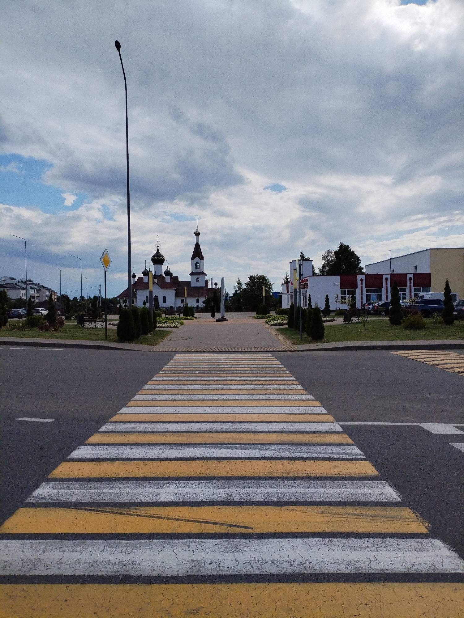 The image captures a street scene in the city of Bialynichy, Belarus, featuring a prominent pedestrian crossing in the foreground. The crossing is marked with alternating yellow and white stripes, clearly indicating a designated area for pedestrians to cross the asphalt road. In the middle ground, a white building with a red roof and dark domes, likely a church, stands tall, suggesting a central or significant location within the city. The church is flanked by manicured bushes and trees, contributing to a well-maintained urban landscape. Beyond the church, other buildings and trees are visible, hinting at the surrounding urban environment. A lamppost stands to the left of the church. The sky is overcast with dramatic clouds, suggesting a slightly moody atmosphere. The image is taken from a low angle, emphasizing the pedestrian crossing and the buildings in the distance.