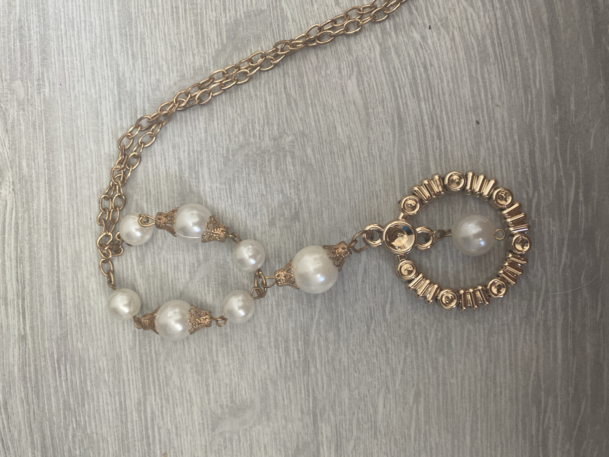 A gold-toned chain necklace with faux pearls and a circular pendant is laid out on a light wood-grain surface. The necklace features a delicate chain, a string of several faux pearls of varying sizes, and a circular pendant adorned with small gold-toned beads and a central faux pearl. The image appears to be a product photograph, taken from directly above, with neutral lighting. There are no people present in the image. The location is not identifiable as Blagoevgrad, Bulgaria.