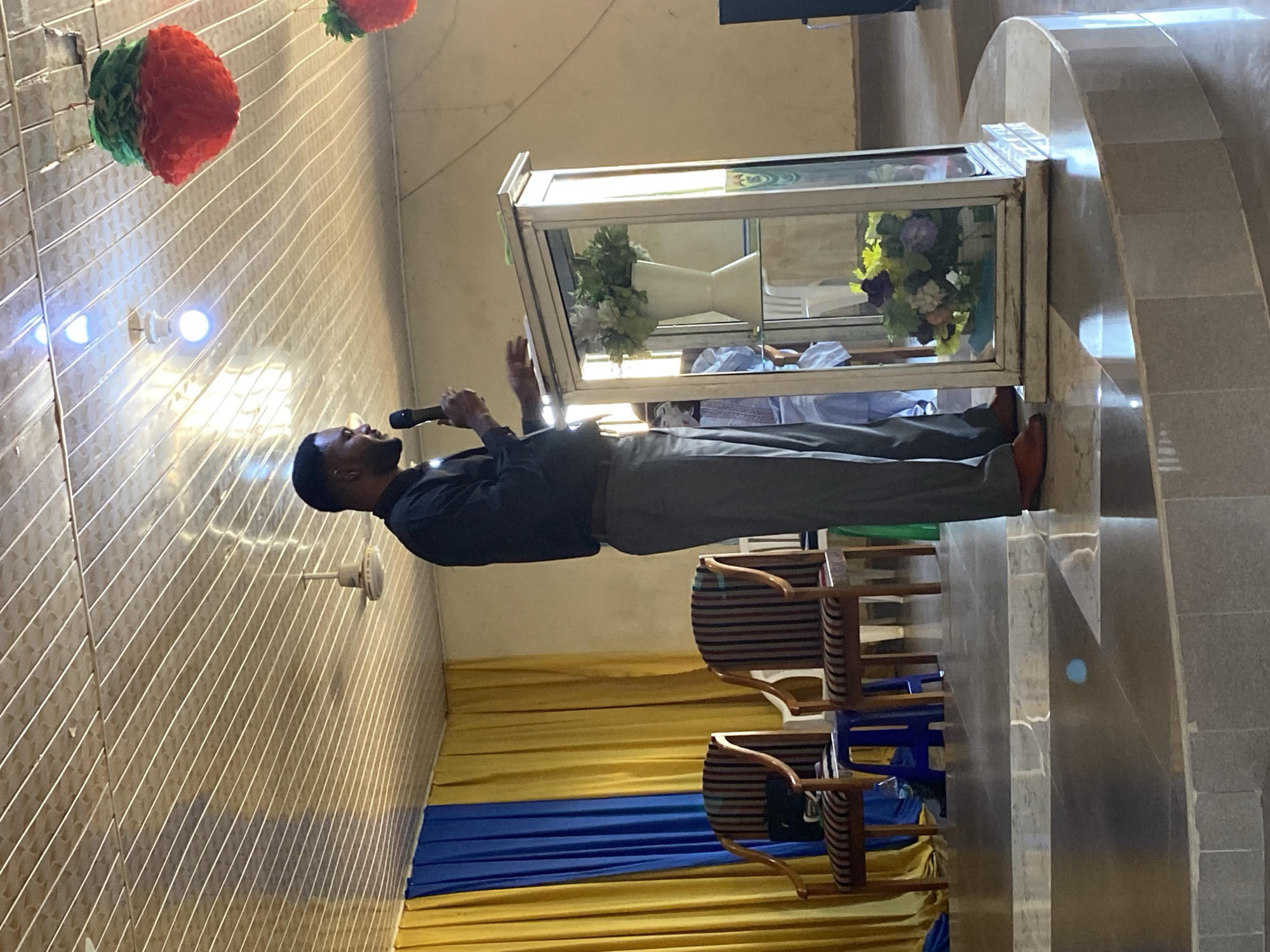 A man is standing and holding a microphone in front of a display case in what appears to be a church or religious gathering place. The room is decorated with yellow and blue fabric drapes, along with red and green decorative balls hanging from the ceiling. Chairs are stacked nearby. The location is in Bukuru, Nigeria.