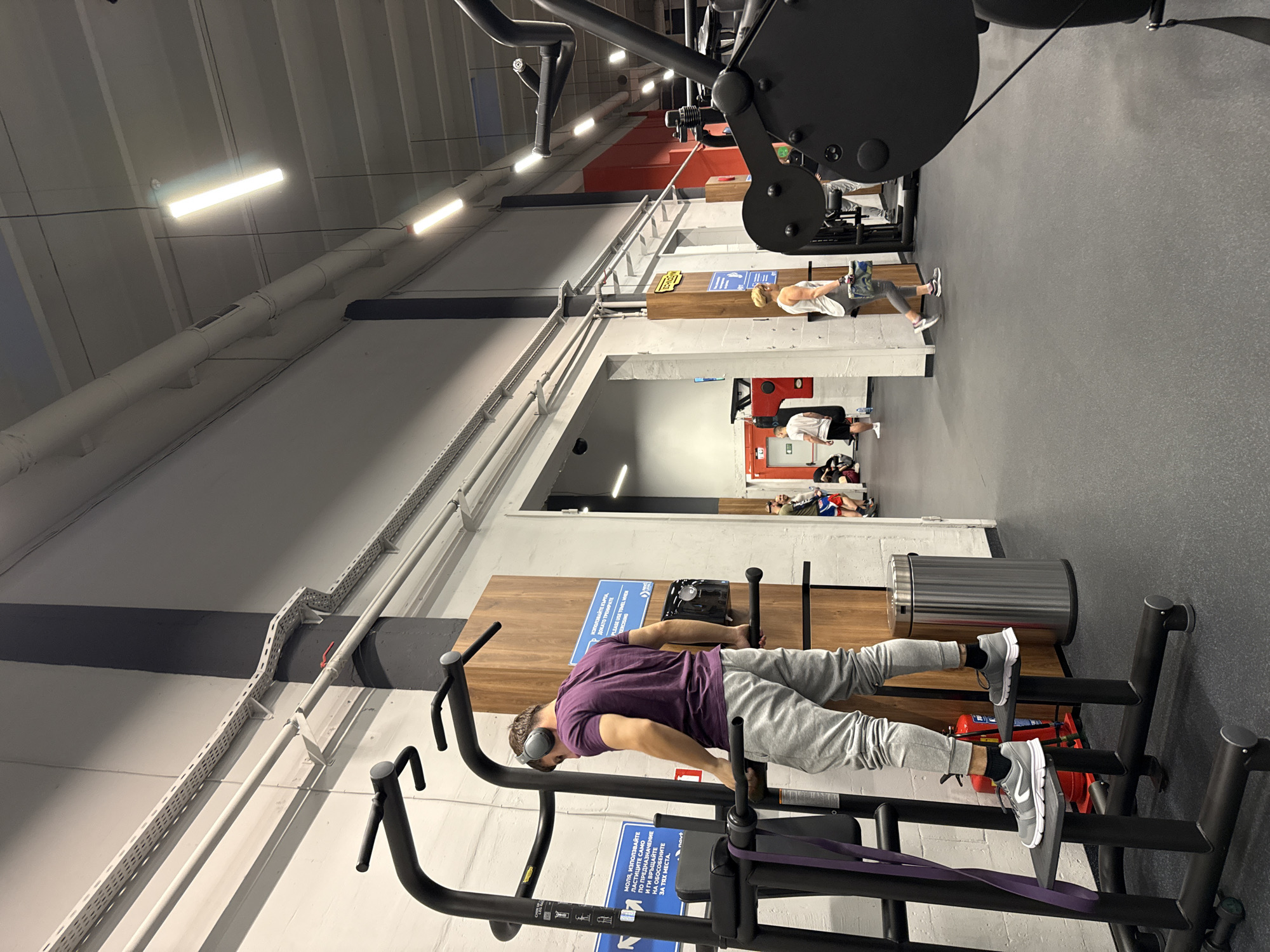 This image captures an indoor gym environment in Sofia, Bulgaria, bustling with activity. The scene is illuminated by bright fluorescent ceiling lights, suggesting it could be any time of day.
In the foreground, a man wearing a purple t-shirt, grey sweatpants, and headphones is performing a bodyweight exercise on a black piece of gym equipment resembling parallel bars. He maintains a horizontal body position, with his legs slightly elevated, indicative of an advanced core and upper body exercise. A purple resistance band is wrapped around the lower part of the equipment near his feet. He appears focused on his workout.
In the mid-ground and background, other gym-goers are visible. One person in a white tank top and grey leggings stands near a wooden structure, possibly stretching or using another piece of equipment. Further back, another individual in a white t-shirt and black shorts is engaged in an activity near a red structure. Several other people are visible in various parts of the gym, with some lying on mats. The floor is a dark grey, typical of gym flooring.
Notable details include industrial-style ceilings with exposed pipes and cable trays. A stainless steel trash can and a red fire extinguisher are visible in the background behind the main subject.
Visible text on blue signs includes instructions in Bulgarian and partially in English. One sign reads: "МОЛЯ, ИЗПОЛЗВАЙТЕ ЛАСТИЦИТЕ САМО ПО ПРЕДНАЗНАЧЕНИЕ И ГИ ВРЪЩАЙТЕ НА ОБОСОБЕНИТЕ ЗА ТЯХ МЕСТА." (PLEASE USE THE BANDS ONLY AS INTENDED AND RETURN THEM TO THEIR DESIGNATED PLACES.), with "novt" branding. Another sign states: "ИЗПОЛЗВАЙТЕ КЪРПА, ДОКАТО ТРЕНИРАТЕ" (USE A TOWEL WHILE TRAINING) and a partial English translation "PLEASE USE TOWEL WHER".
