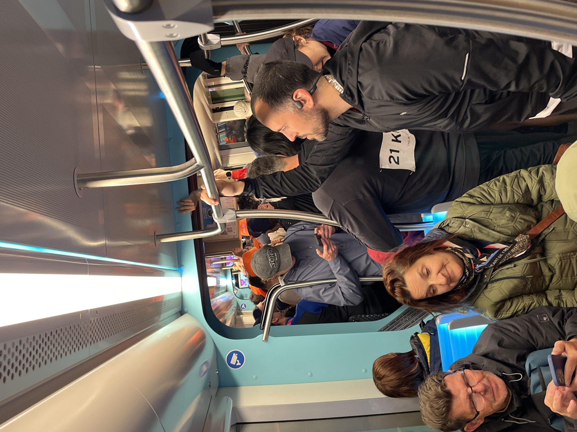 The image depicts a crowded train carriage, likely in Luxembourg City, Luxembourg, based on the design of the carriage and the presence of individuals. The passengers are standing and holding onto the overhead handrails, with multiple individuals visible. A man with a visible '21K' bib is present. The interior of the train carriage is modern, featuring blue and white paneling.