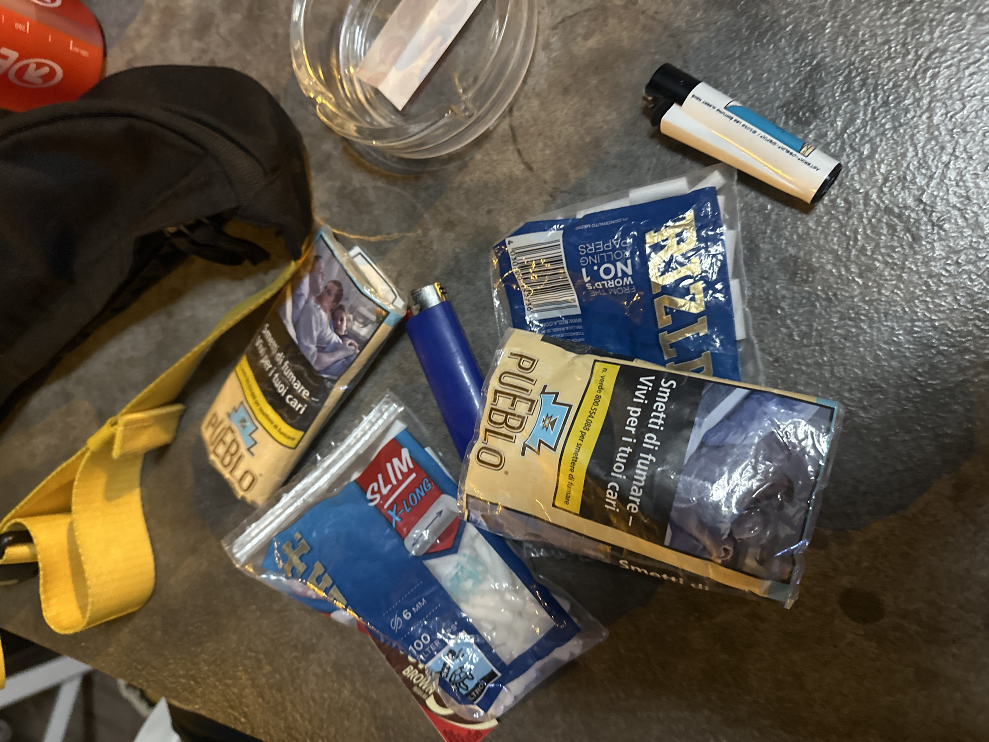 The image depicts a collection of smoking-related items scattered on a textured, dark gray surface, possibly a table or countertop, which appears to be outdoors. In the background, a yellow strap is visible, and a dark fabric bag is partially seen. A red cylindrical object, possibly a container, is also in the background.
The foreground is dominated by several packets of rolling papers and filters. One packet, prominently displayed, is yellow with "PUEBLO" written in large letters. It also shows text in Italian, "Smetti di fumare - Vivi per i tuoi cari" (Stop smoking - Live for your loved ones), along with a health warning image of a person. Another blue packet, also with "PUEBLO" branding, is labeled "WORLD'S NO.1 ROLLING PAPERS". A packet of "SLIM X-LONG" filters, with "100 FILTERS" and a diameter of "6 MM", is also visible, as is a packet of "BROWN" rolling papers.
A blue lighter lies next to the packets, and a clear glass ashtray with a single white rolling paper inside is situated to the right. A white and black disposable lighter is also present in the lower right corner. The overall scene suggests a casual setting where these items are laid out, potentially before or after use.
The specific location in the city of Ospedaletti, Italy, cannot be determined from the image alone.