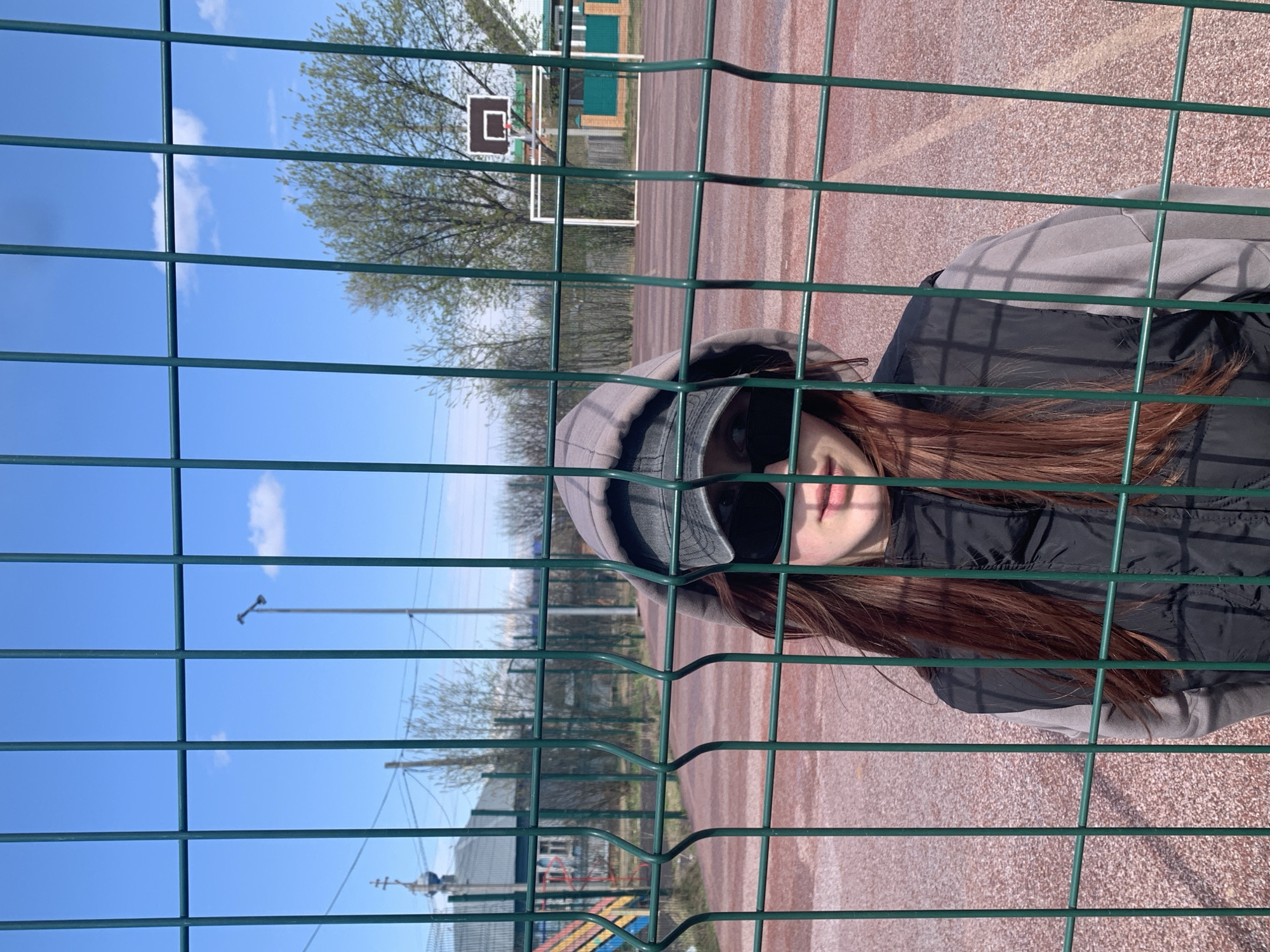 The image captures a person looking through a dark green wire mesh fence on a bright, sunny day at an outdoor sports facility. The subject, visible on the right side of the frame, wears a grey hooded sweatshirt, a black padded vest, and a black cap, partially obscuring their face. Long brown hair flows around their shoulders. Their lips are slightly parted, and they appear to be looking directly at the camera.
The fence forms a prominent grid pattern across the foreground. Behind it, a reddish-brown, speckled surface, resembling an asphalt or tartan sports court, stretches into the background. Various sports equipment is visible, including a basketball hoop with a white backboard and black rim, and further back, a white-framed soccer goal.
In the midground, several trees with early spring foliage or bare branches are scattered. A small building with a green roof can be seen beyond the court. The background is dominated by a clear blue sky with a few sparse white clouds. Distant power lines, poles, and a faint structure resembling a church dome are also discernible. The setting suggests a community or school sports ground in Фоки, Russia, under bright daylight. No visible text is present in the image.