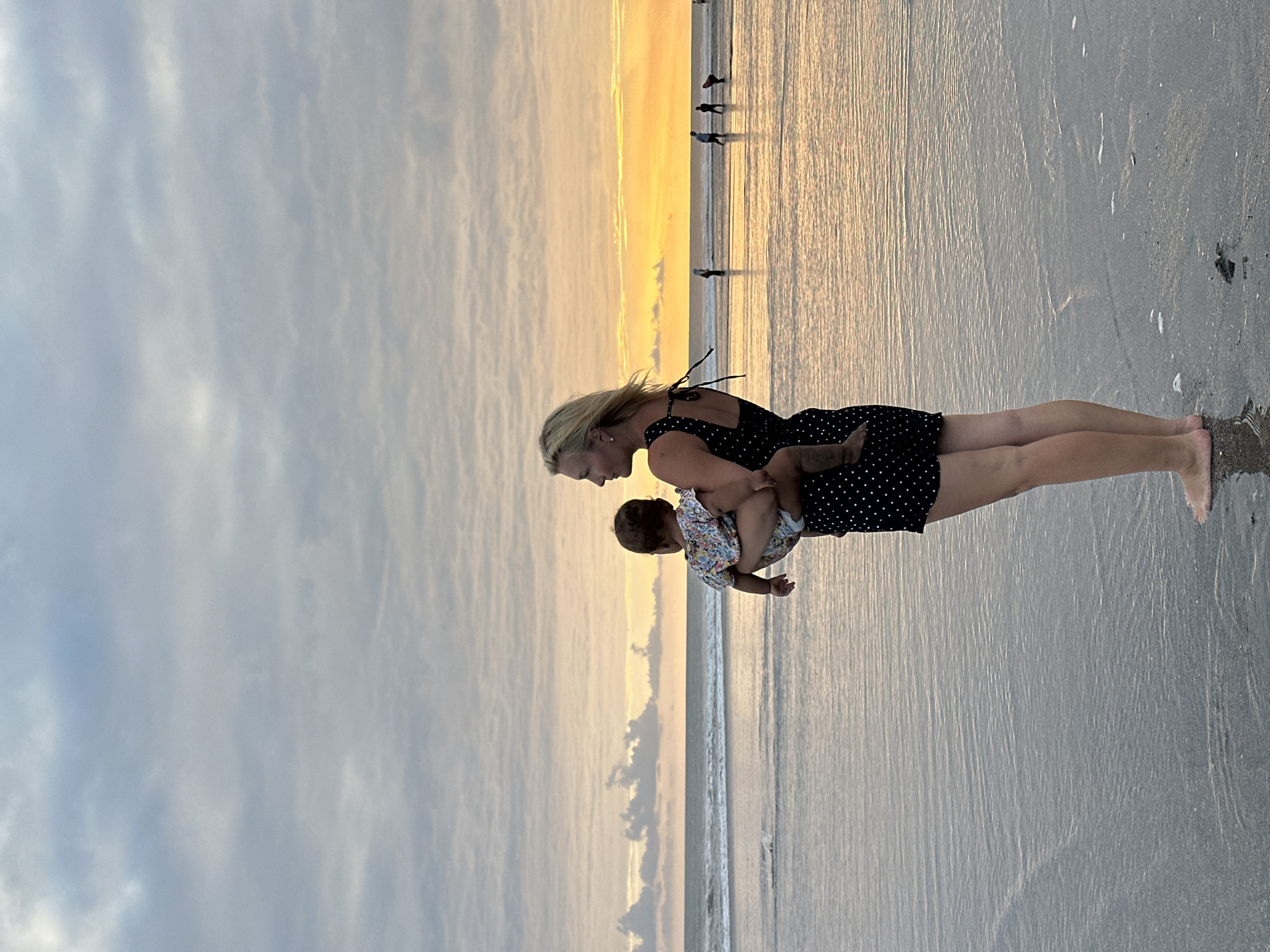 The image depicts a serene beach scene at dusk, with a woman holding a baby. The woman, with blonde hair, is wearing a black polka-dot dress and is facing downwards, looking at the baby she holds. The baby, wearing a floral-patterned outfit, is facing away from the camera, their gaze seemingly directed towards the ocean. The beach is wet, with shallow water lapping at the shore, reflecting the warm hues of the setting sun.
In the background, silhouetted figures of people are visible walking along the shoreline, suggesting a public beach. The sky is filled with soft, layered clouds, illuminated by the golden light of the sunset, creating a peaceful and tranquil atmosphere. The water is calm, with gentle waves breaking on the sand. The location is identified as North Kuta, Indonesia. The time of day is clearly sunset, indicated by the warm, soft light and the color of the sky. The weather appears to be clear and mild, suitable for a beach outing. The overall mood conveyed by the image is one of tenderness and calm, capturing a quiet moment between a mother and child against the backdrop of a beautiful sunset. There is no visible text in the image.