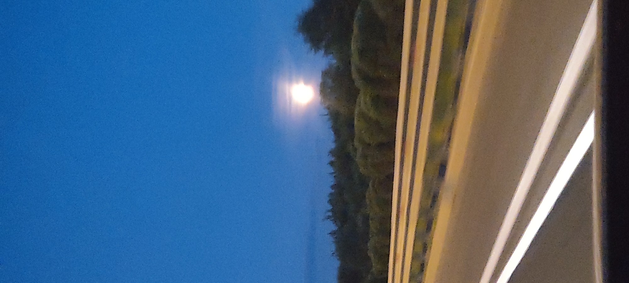 This is a vertical, slightly grainy photograph of a night sky with a bright moon visible. The sky is a deep, clear blue, indicative of twilight or early night. To the right of the sky, there is a dark silhouette of trees, beyond which are what appear to be lights from a road or a building. The road itself is paved with visible lane markings that are illuminated, suggesting artificial lighting. The overall impression is of a serene, possibly rural or suburban landscape at dusk or night. There is no discernible text in the image.