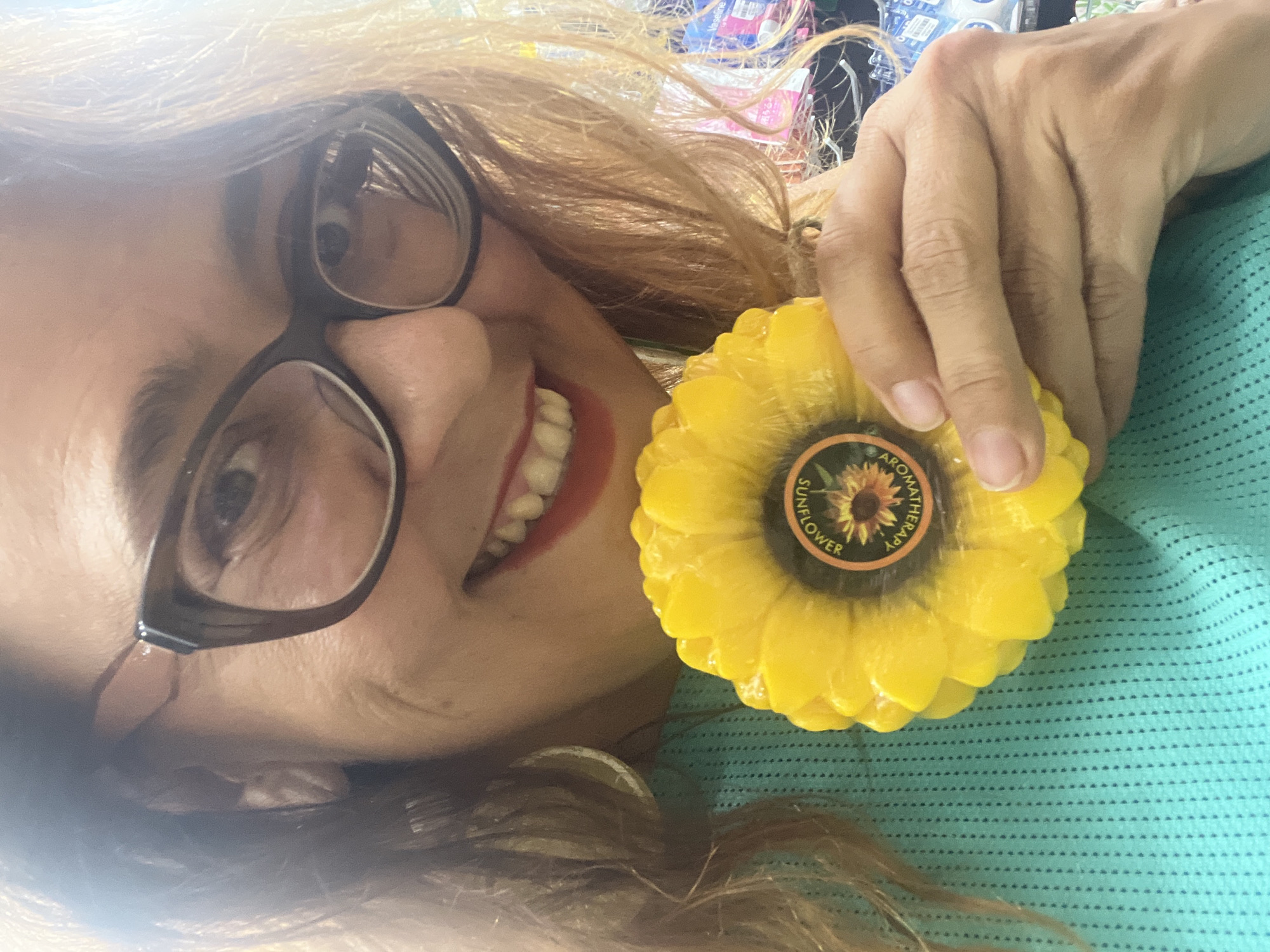 A close-up, waist-up, selfie-style shot captures a woman holding a sunflower-shaped object with a label that reads "SUNFLOWER AROMATHERAPY." The woman is wearing glasses and a teal, mesh-like top. Her hair is light brown and frames her face. The background is blurred, but appears to be an indoor setting with shelves stocked with various products. The lighting is bright, causing some glare on the woman's face and glasses. The image was taken in Nha Trang Ward, Vietnam.