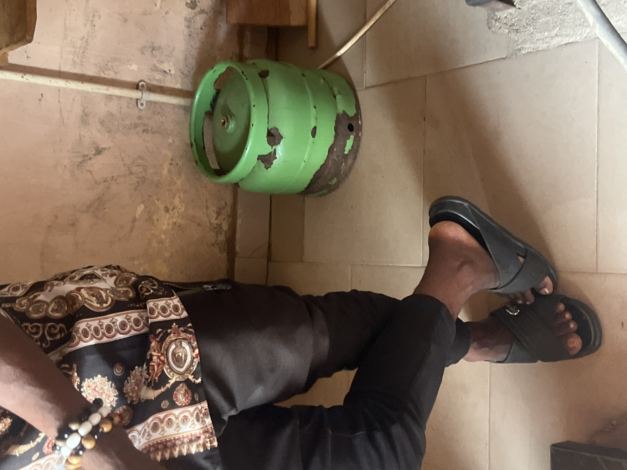 The image shows a person's legs and torso, with a green gas cylinder in the background. The person is wearing black pants and a black and gold patterned shirt. They are also wearing black sandals. The gas cylinder appears to be old and rusted, with some of the green paint chipped off. The scene appears to be indoors, with a tiled floor and a beige wall. There is a white pipe running along the wall, attached by metal clips. The lighting suggests it is daytime. The overall impression is of a casual, everyday scene, possibly in a home or workshop. The location is identified as Jalingo, Nigeria. No text is visible in the image.