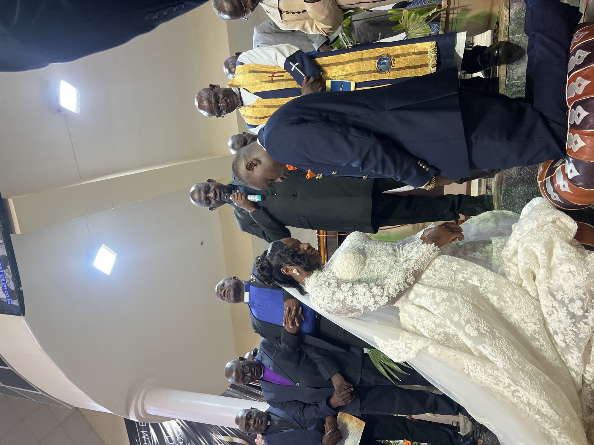 The image depicts a wedding ceremony taking place indoors, likely in a church in Kabong, Nigeria. In the foreground, a bride in a white lace wedding gown with a long veil is kneeling, her head bowed. Beside her, the groom, wearing a dark blue suit with an orange boutonnière, is also kneeling. Several men in clerical attire stand behind them, observing the ceremony. One man in the center, wearing a black suit and purple collar, is holding a microphone and appears to be officiating. Another man, wearing a gold and blue stole over his clerical attire, also stands nearby. The background shows architectural elements of the building, including a curved ceiling with lighting fixtures and a banner with text that is partially visible. The lighting suggests an indoor setting, possibly a church auditorium. The overall atmosphere appears solemn and ceremonial.