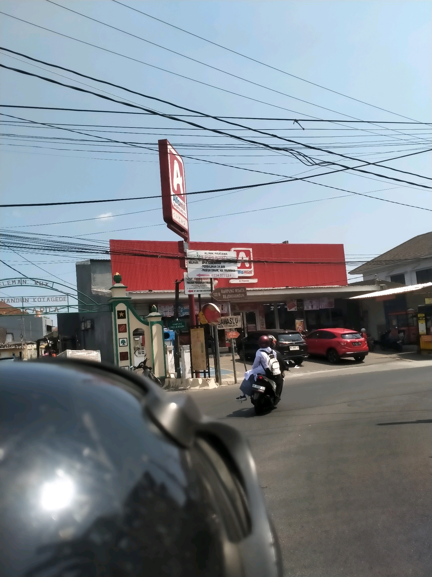 The image captures a street scene in Yogyakarta, Indonesia, likely near the historic Kotagede area, as suggested by signage. Dominating the frame is a red-roofed Alfamart convenience store, its bright facade contrasting with the blue sky overhead, crisscrossed by a dense network of electrical wires. A tall, red sign with a white "A" logo, a signature of Alfamart, stands prominently to the left of the building.
In front of the store, a black SUV is parked, and to its right, a red car is also visible. A motorcycle carrying two individuals is in motion on the asphalt road, heading away from the camera. The rider, wearing a white shirt and backpack, is in the foreground of the motorcycle. The environment appears to be a commercial district with the presence of shops and a gateway with decorative pillars. The overall impression is a typical urban street view in Indonesia, characterized by its infrastructure and daily life activities.