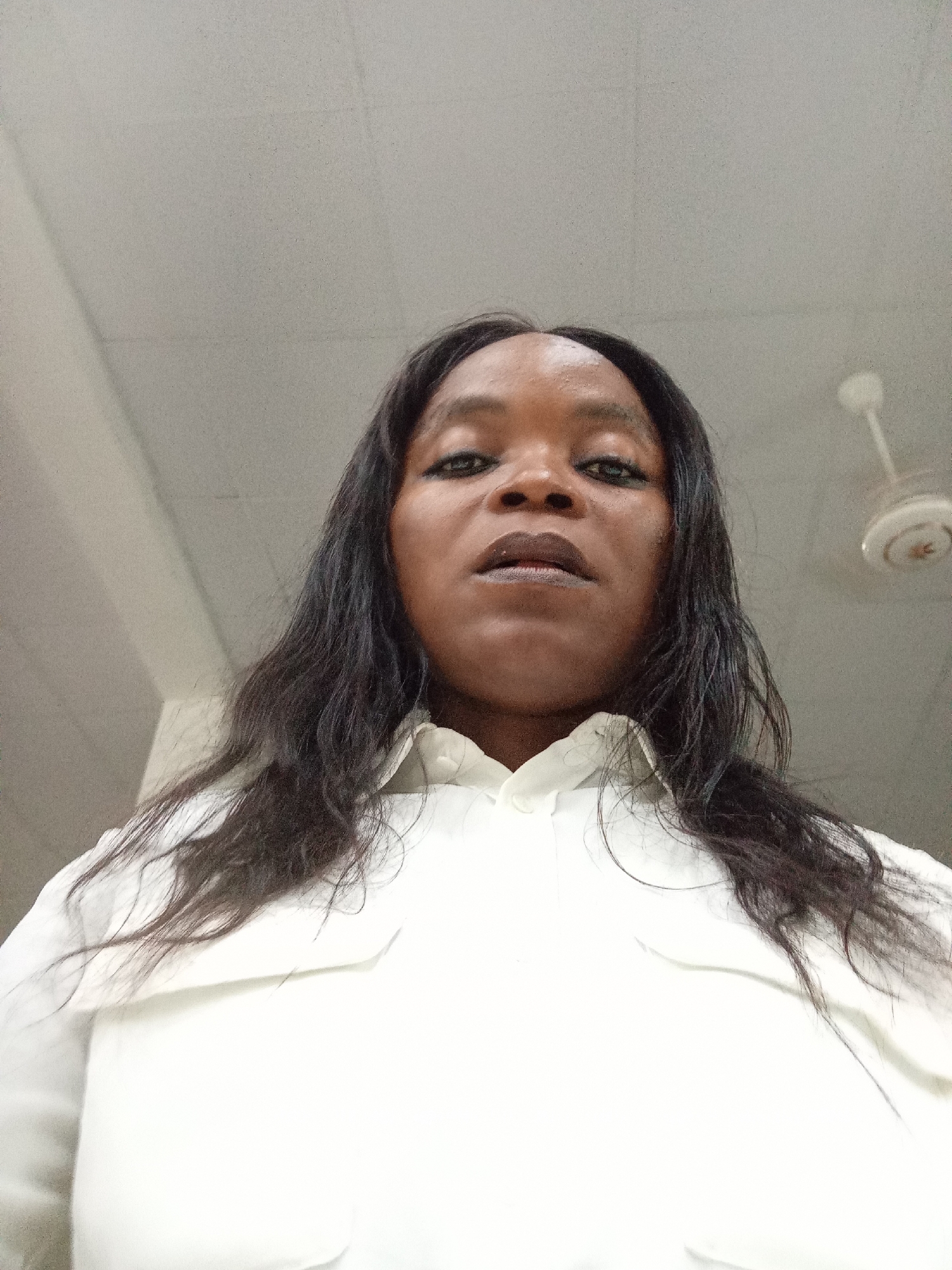 A low-angle, head-on, full-face selfie taken from a woman's perspective is presented. The woman has dark, straight hair that extends past her shoulders. She is wearing a white collared shirt. The background consists of a white ceiling with what appears to be a ventilation fan mounted on it. The lighting suggests an indoor environment. The image does not provide any contextual information to determine its location in Kason, Nigeria.