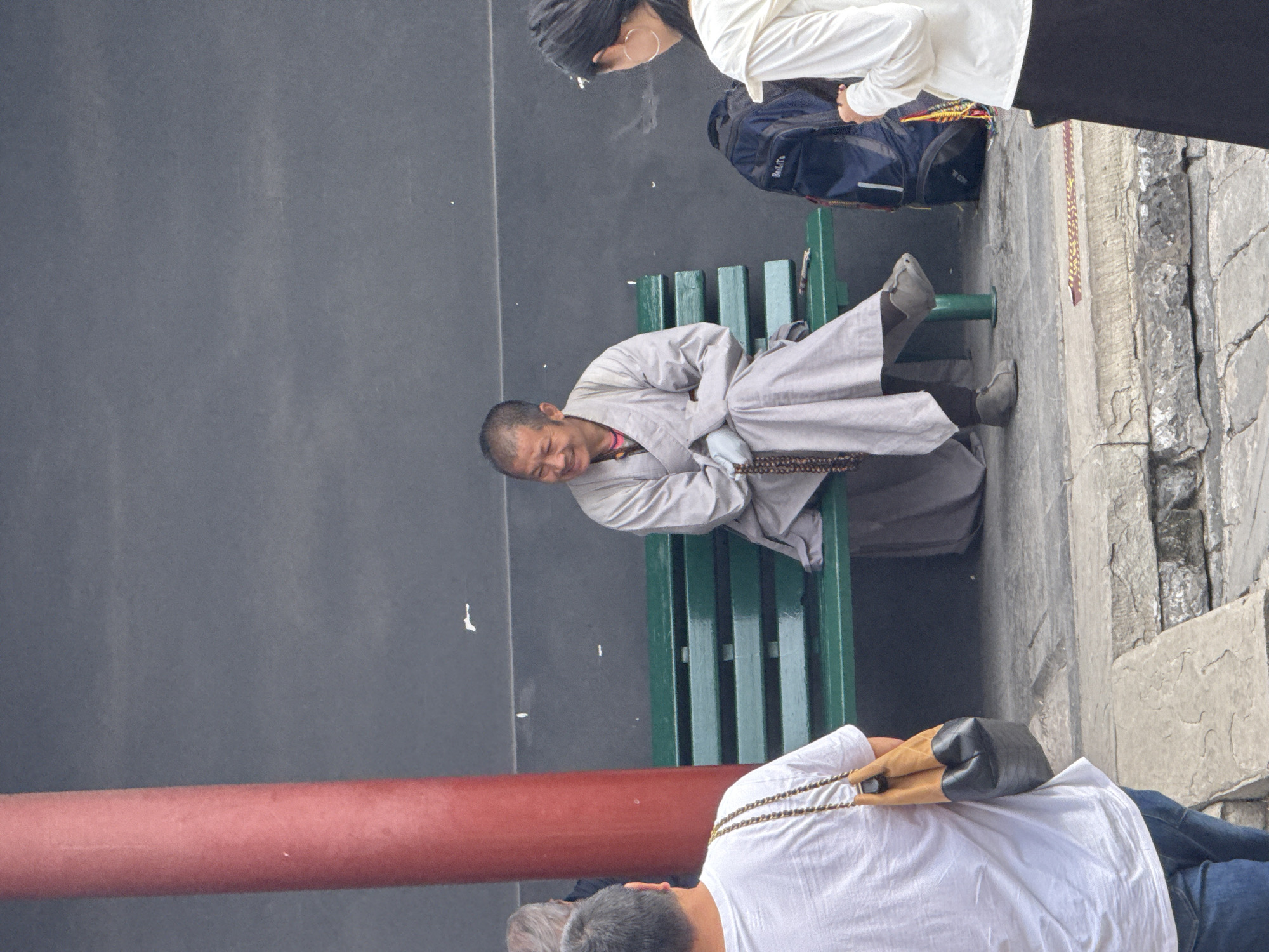 The image depicts a street scene with several individuals. In the center, a person wearing a light grey robe and beads around their neck sits on a green bench, smiling. To the left, a person in a white t-shirt is seen from the back, carrying a brown and black bag. To the right, a person in a white top and dark skirt stands, facing away from the viewer. A blue backpack is on the ground next to the person in the white top. A thick red pillar is visible on the left side of the frame. The background consists of a dark grey wall. The ground is paved with stone. This scene is located in Dongcheng District, China.