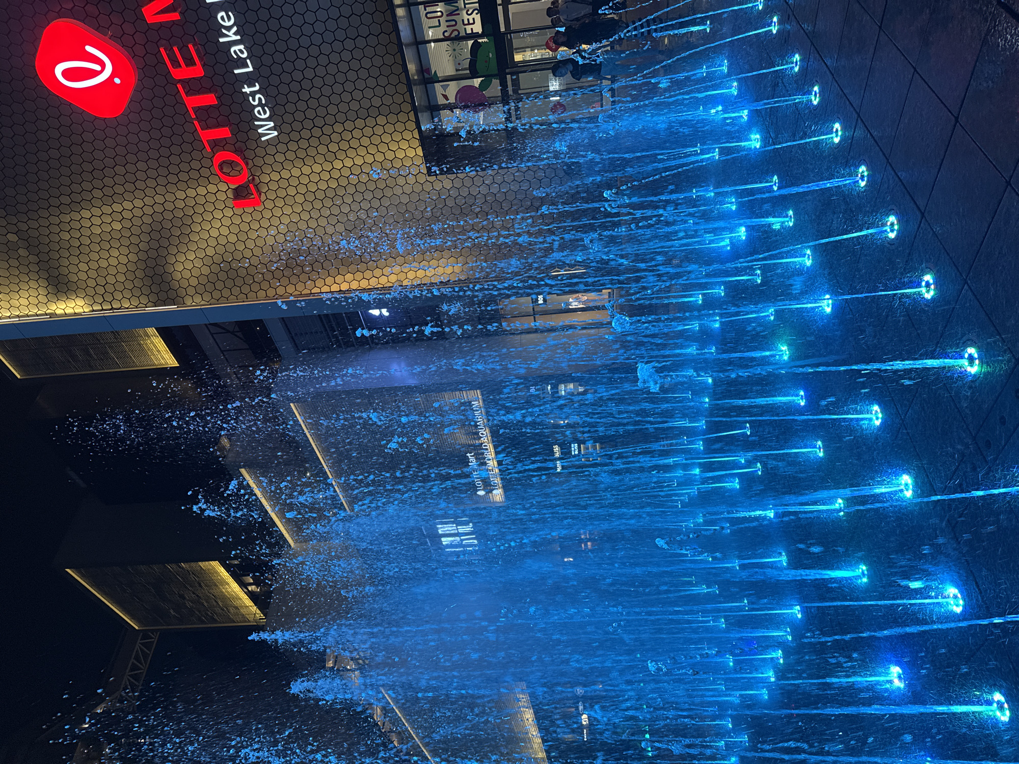 The image captures an illuminated water fountain display in front of a modern building at night. The fountain features numerous streams of water, lit from below with vibrant blue and some green lights, creating a dynamic and colorful spectacle. In the background, the building's facade is visible, adorned with a large red logo and text that reads "LOTTE MART" and "LOTTE WORLD AQUARIUM". Further text on the building includes "West Lake Ha Noi". The architecture is contemporary, with honeycomb-like patterns on the building's exterior and illuminated accent lighting. A few people are visible in the background, suggesting a public or commercial space. The overall scene conveys an atmosphere of urban entertainment and nighttime ambiance in Hanoi, Vietnam.