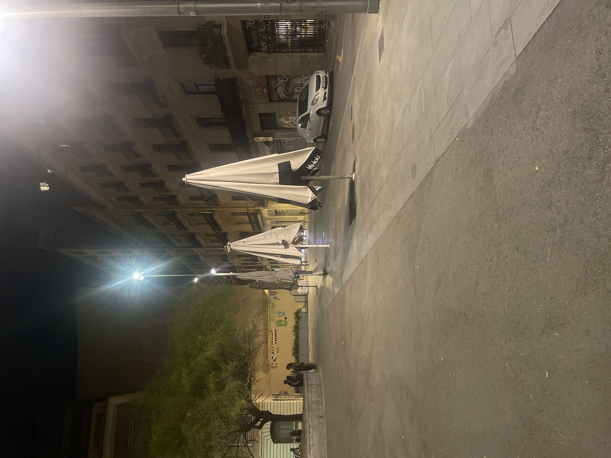 The image depicts a street scene at night in Barcelona, Spain, specifically on Carrer dels Tallers. A bright streetlamp illuminates the area, creating lens flare. On the left, a large tree with green foliage is situated next to a low, curved stone wall with people sitting on it. Further left, a building with a pale facade and windows is visible. The center of the image features three large, closed white umbrellas, likely from outdoor seating for a cafe or restaurant. The umbrellas are positioned on a paved walkway. In the background, an apartment building with multiple floors and balconies stretches along the street. A white car is parked in front of a building with graffiti on its facade. The street is made of asphalt and stone pavement. The overall atmosphere is quiet and deserted, suggesting late hours or off-season.