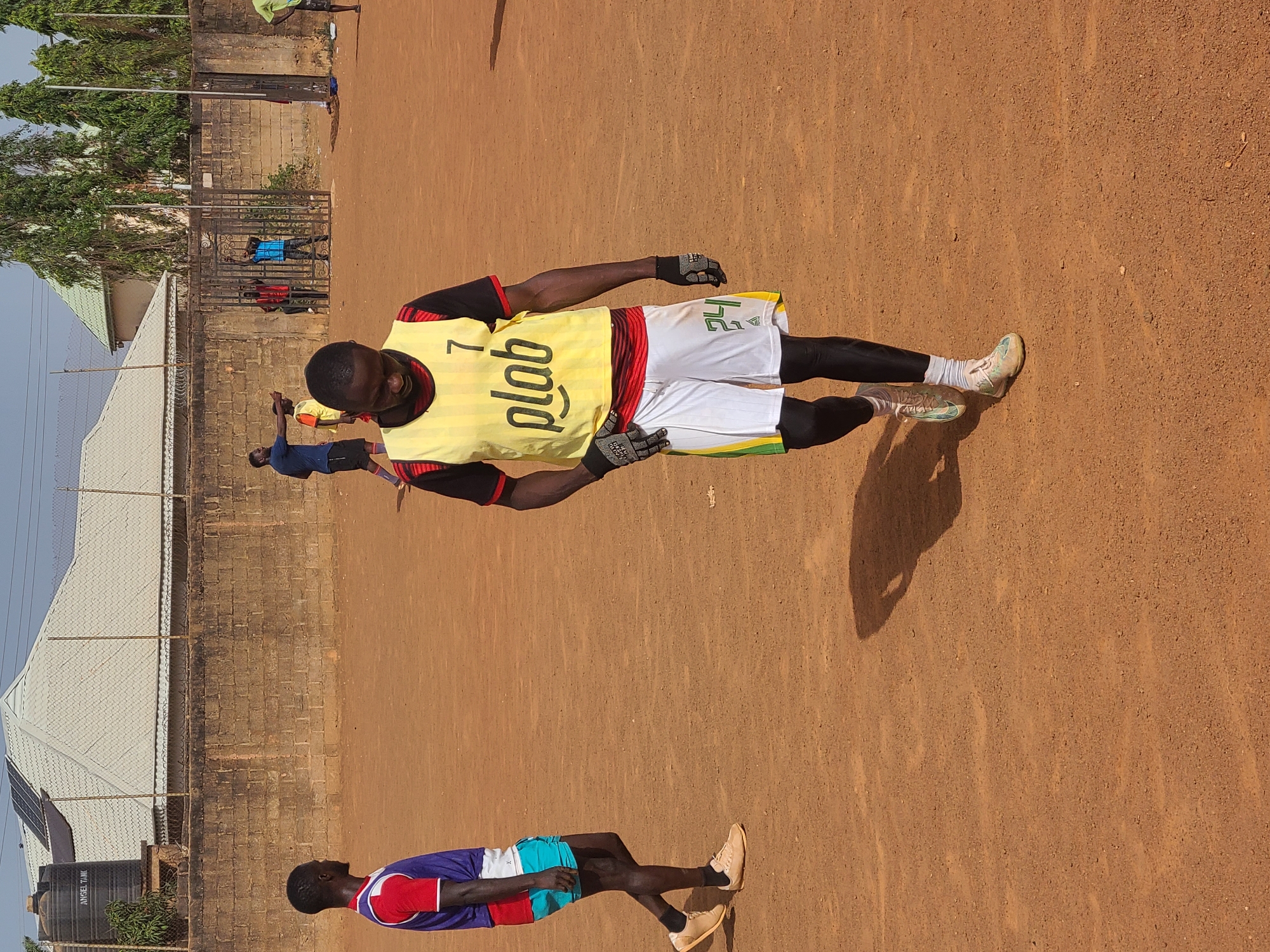 The image shows a dusty field in Barakin Lerawi, Nigeria, where several people are present. One man is wearing a yellow jersey with "plab" printed on it, along with white shorts and gloves, and is standing on the field. Another person is wearing a blue and red shirt with turquoise shorts and is also standing. Other individuals are visible in the background, and there is a fence and buildings surrounding the field.