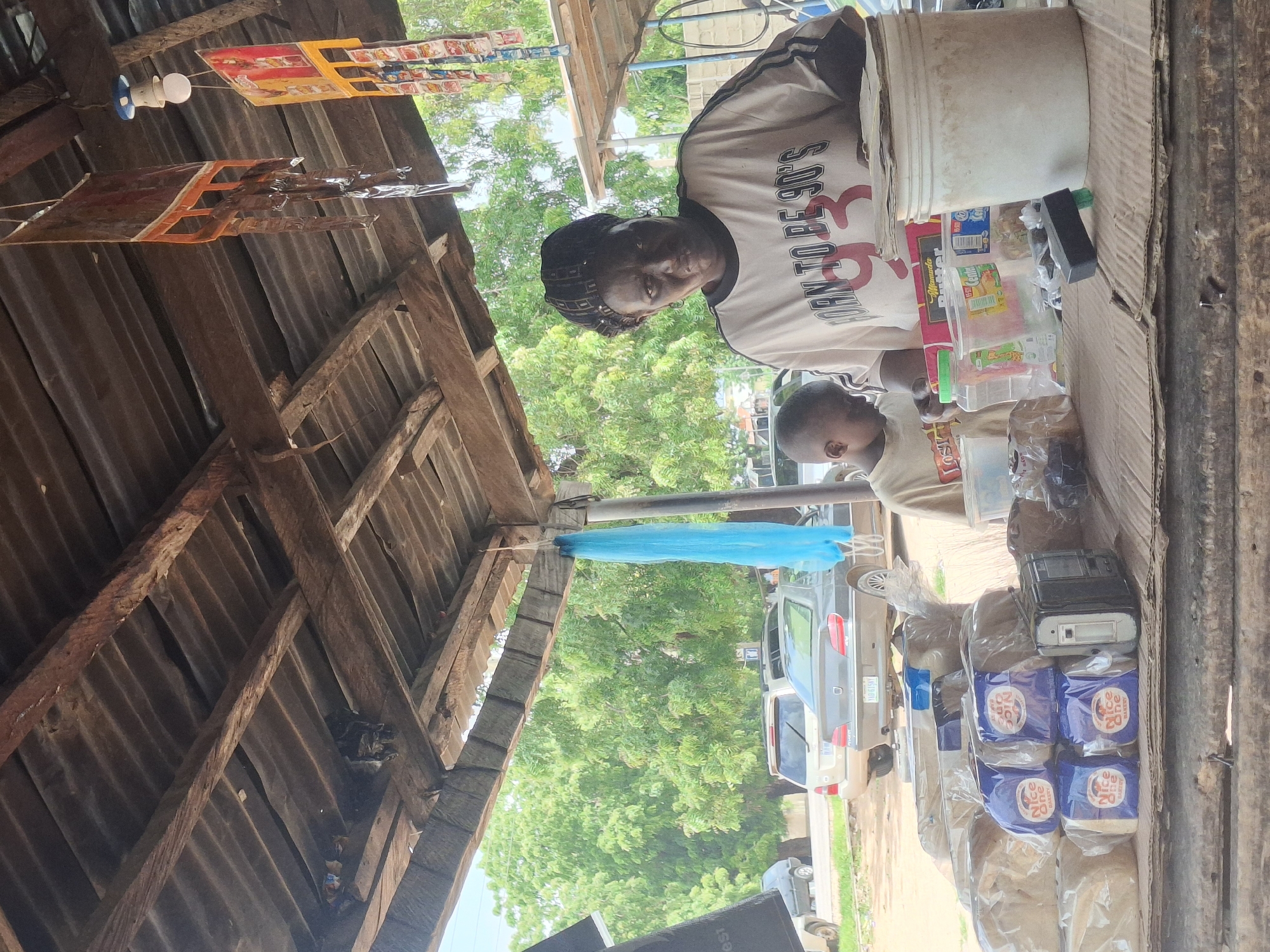 This image depicts a scene at a roadside stall in Biliri, Nigeria. A woman, appearing to be the vendor, stands behind a wooden counter laden with products. She is wearing a light-colored t-shirt with "BORN TO BE 90'S 93" printed on it. A young boy, presumably a customer or helper, stands to her left, looking down.
On the counter, there are several loaves of bread wrapped in clear plastic, with some bearing the "Nice One Bakery" logo. To the right of the bread, a black and silver electronic device, possibly a radio or recorder, is visible. Several containers and packages, including what appears to be butter and other food items, are also present. A large white bucket sits on the right side of the counter.
The stall itself is constructed with a corrugated metal roof supported by wooden beams and posts, suggesting a simple, open-air setup. Behind the stall, trees and vegetation are visible, and several cars are parked on a road. The overall impression is of a bustling local marketplace.