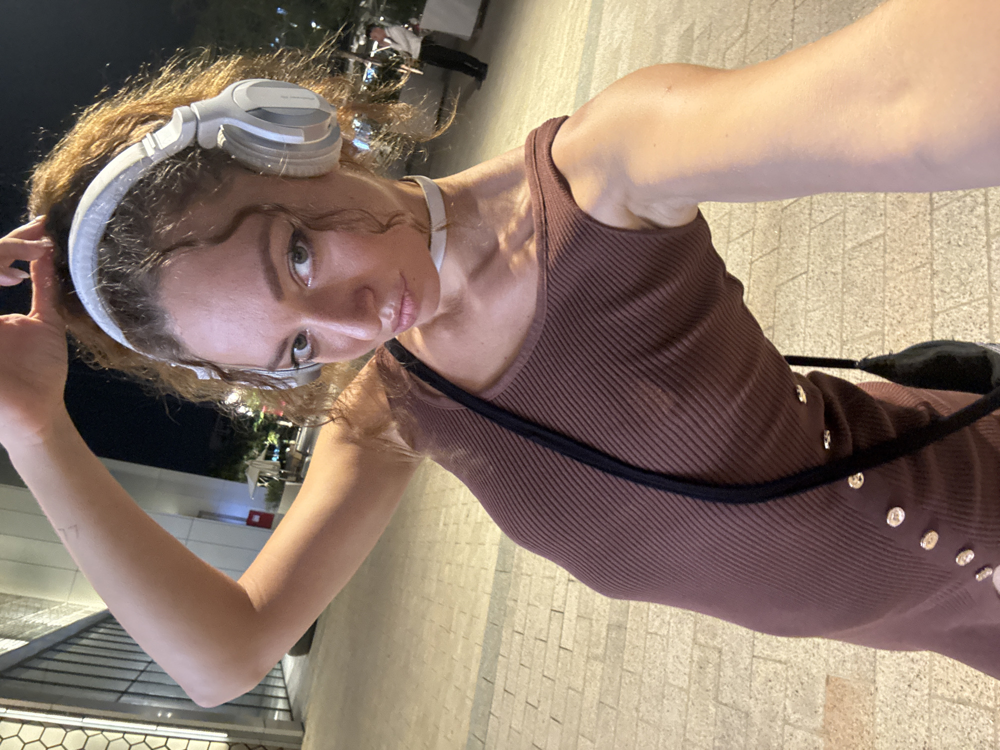 A woman with curly brown hair is taking a selfie at night. She is wearing white headphones and a brown off-the-shoulder top with buttons down the side. A black strap from a bag is visible across her chest. She is standing on a paved walkway in a city, with buildings and trees in the background. The lighting suggests it is an urban area at night. There is a hint of a tattoo on her left forearm. In the background, a man in a white shirt and black pants is visible. The scene does not contain any specific elements that would indicate it is located in Hà Nội, Vietnam.