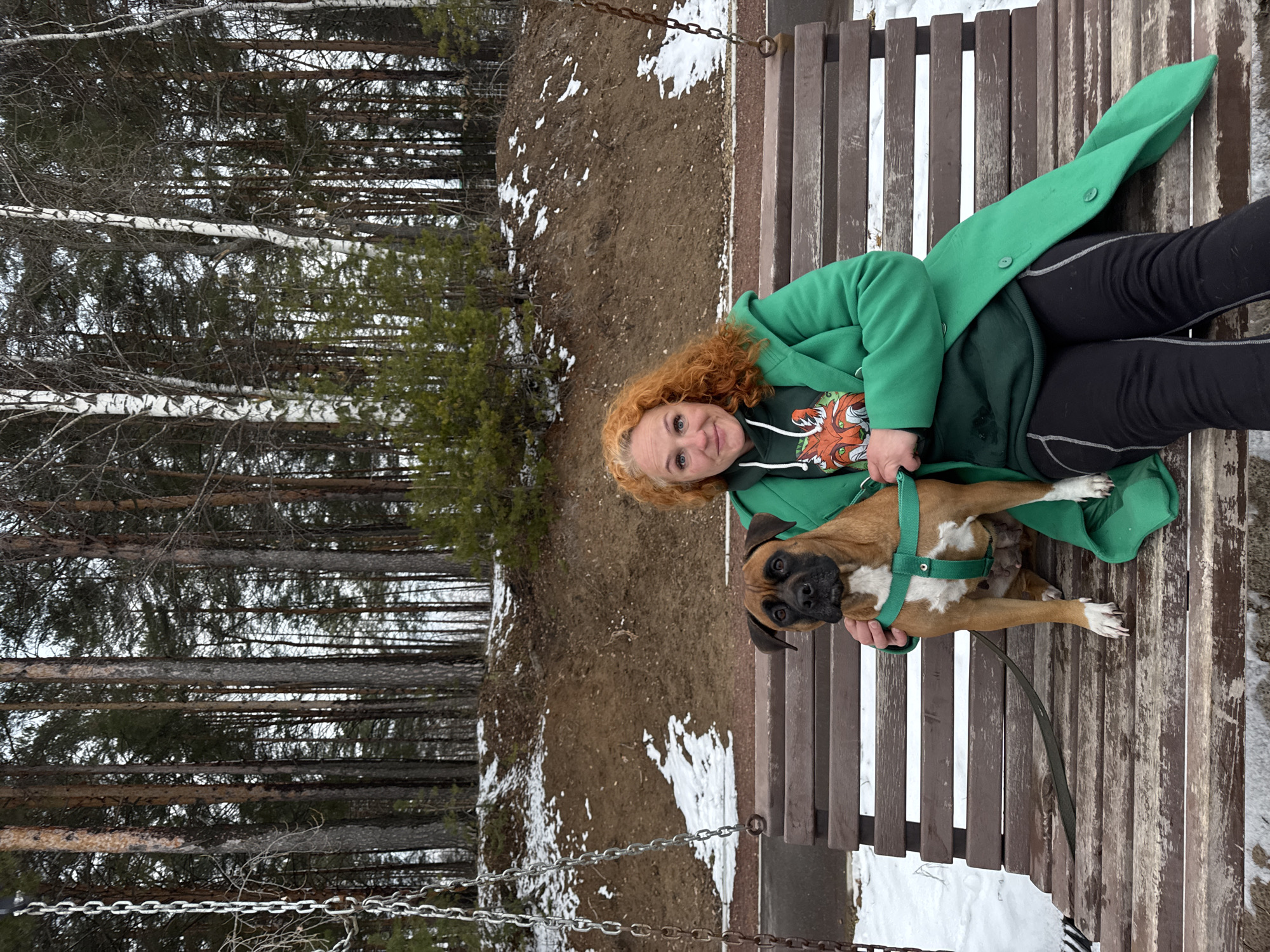 The image shows a woman with curly red hair sitting on a wooden swing bench with a brown and white boxer dog in her lap. She is wearing a green hooded sweatshirt with an orange design and a green coat, along with black leggings with white details. The dog is wearing a green harness and leash. The background includes a forest with bare trees and patches of snow on the ground. The scene appears to be outdoors, possibly in a park. The location is the city of Chaikovsky, Russia.