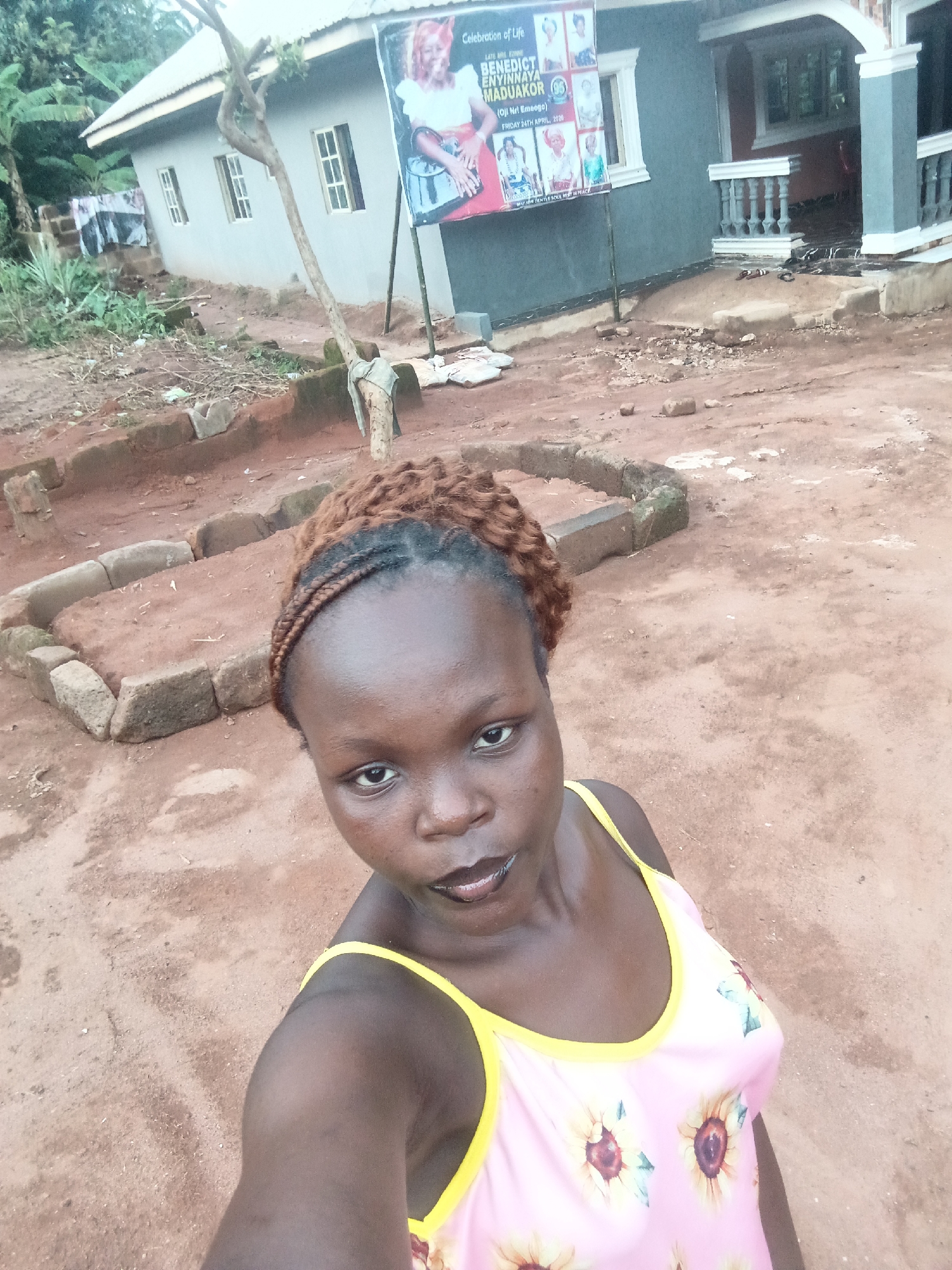 A young woman with dark skin and braided, reddish-brown hair takes a selfie, looking directly at the camera with a neutral expression. She is wearing a pink tank top with yellow trim and sunflower patterns.
The outdoor setting features reddish-brown, unpaved ground, typical of rural or semi-urban areas like Eziudo, Nigeria. In the foreground and mid-ground, there are concrete blocks forming low rectangular borders on the ground, and a slender tree trunk extends upwards.
In the background, two houses are visible. To the left, a single-story house with a light grey exterior, white-framed windows, and a corrugated metal roof is partly obscured by tropical foliage. To the right, another house, painted grey and white with decorative white trim and pillars, features a covered entrance.
Prominently displayed between the woman and the right-side house is a large vertical banner on poles. The banner is a "Celebration of Life" announcement for "LATE MRS. EMMANUEL BENEDICT ENYINNAYA MADUAKOR (95)". It includes the location "(Oji Nri Emeoga)" and the date "FRIDAY 24TH APRIL, 2020," ending with the phrase "MAY HIS GENTLE SOUL REST IN PEACE." The banner features several images, including a large portrait of a woman. The scene is illuminated by diffused daylight, suggesting an overcast day.