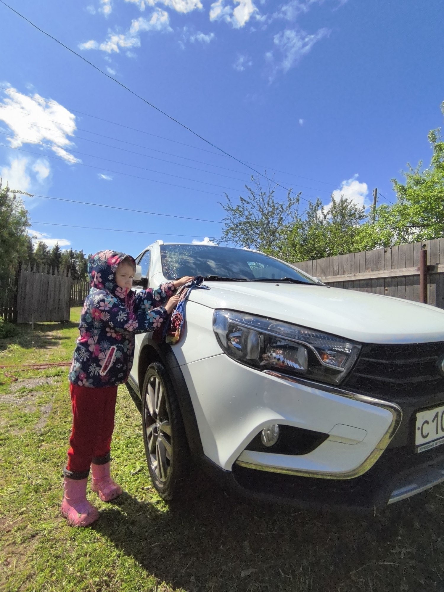 A young child in a floral jacket, red pants, and pink rubber boots is washing a white car on a grassy lawn. The car is positioned near a wooden fence with trees visible in the background. The sky is blue with scattered white clouds. The location appears to be a residential area. The car has a license plate with characters "C 10" visible.